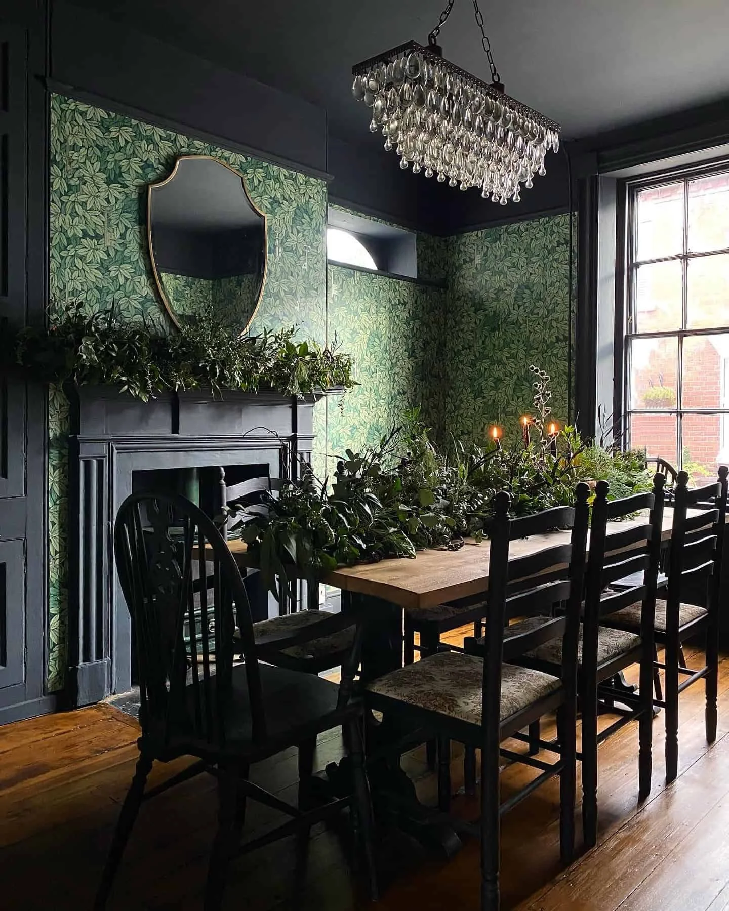 A dining room with a wooden table surrounded by black chairs, decorated with greenery and candles, green leafy wallpaper, a large window, a black fireplace with a mirror above, and a chandelier hanging from the ceiling, moody interior colours