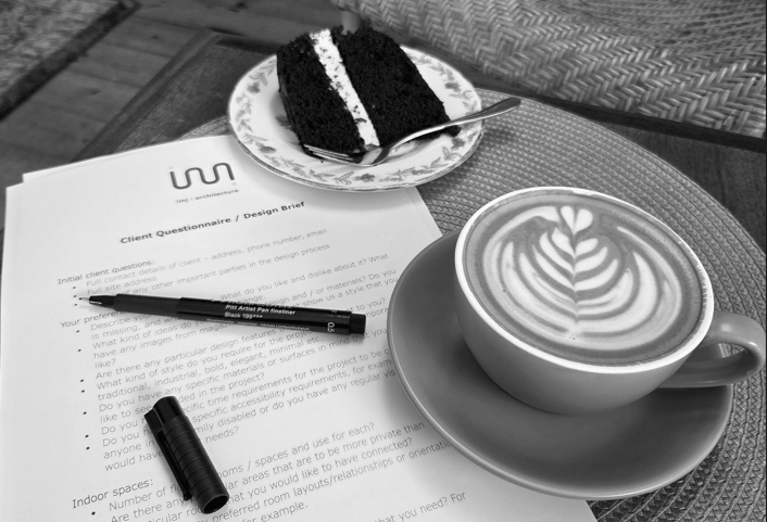 Architectural designer client meeting with a slice of black chocolate cake, a cup of coffee, a client questionnaire and design brief document, and a black marker on a textured table surface.