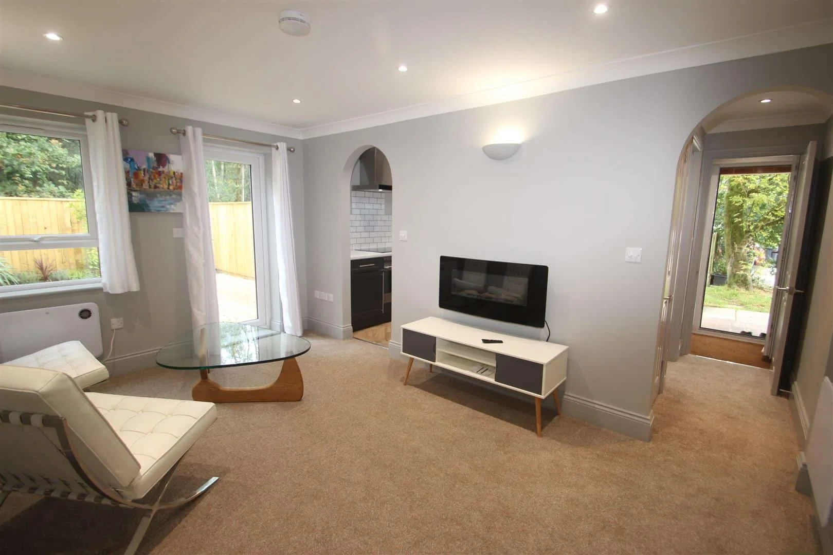 Living room with beige carpet, white furniture, glass coffee table, mounted flat-screen TV, and large windows with white curtains overlooking a backyard with trees and a wooden fence.