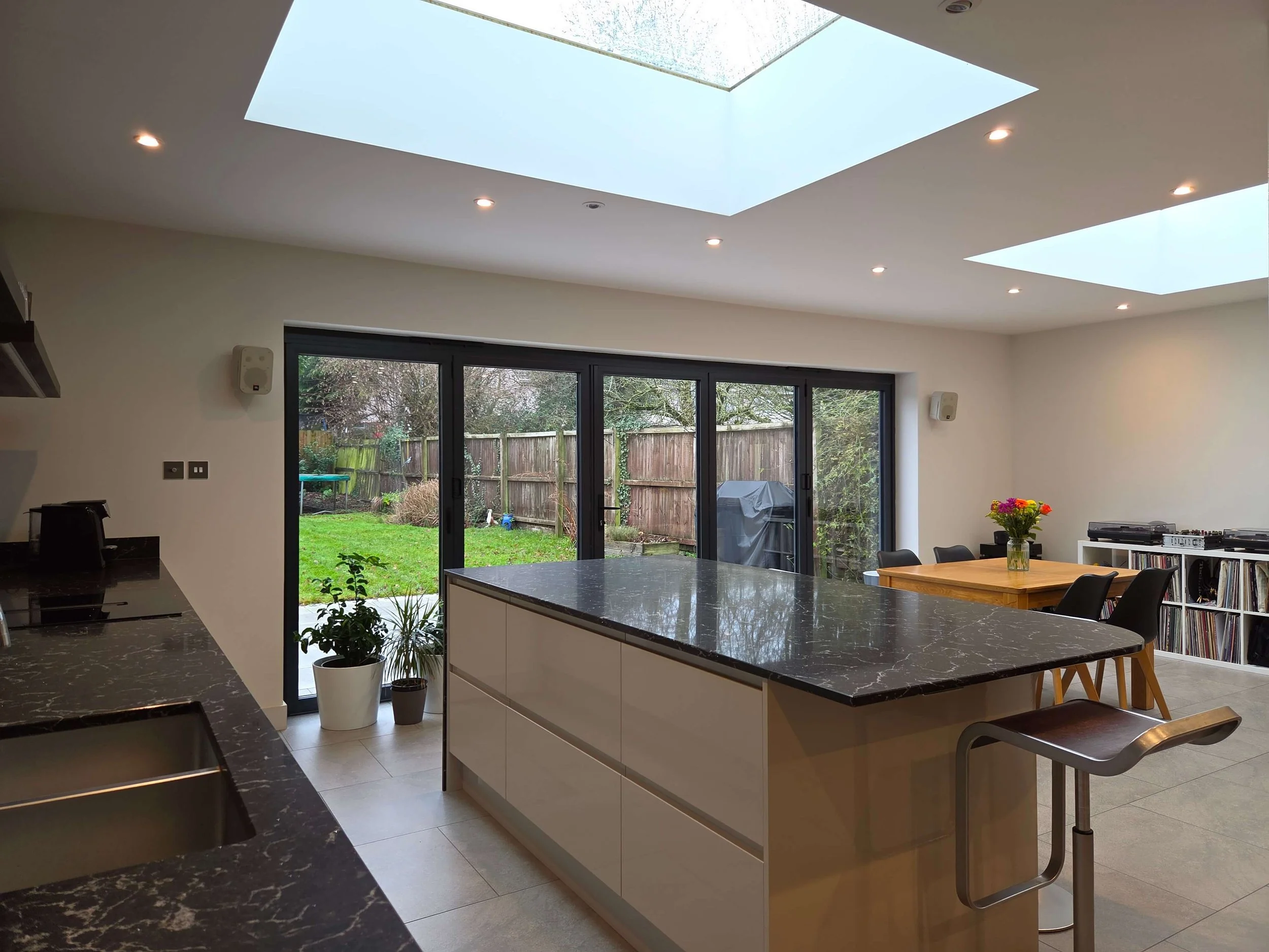 Modern kitchen with a large kitchen island and marble countertop, overlooking a rear garden through large glass sliding doors, with a seating area and potted plants.
