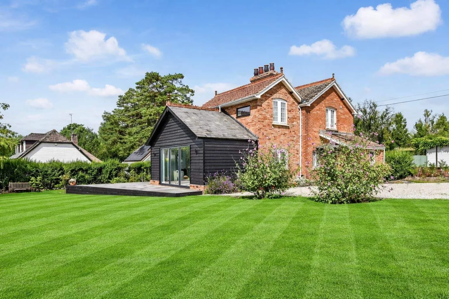 Traditional rural cottage with chimney, a two storey brick extension, single storey timber weather boarded extension, bi-fold glazed doors, surrounded by a private lawn and garden with bushes, under a blue sky with clouds.