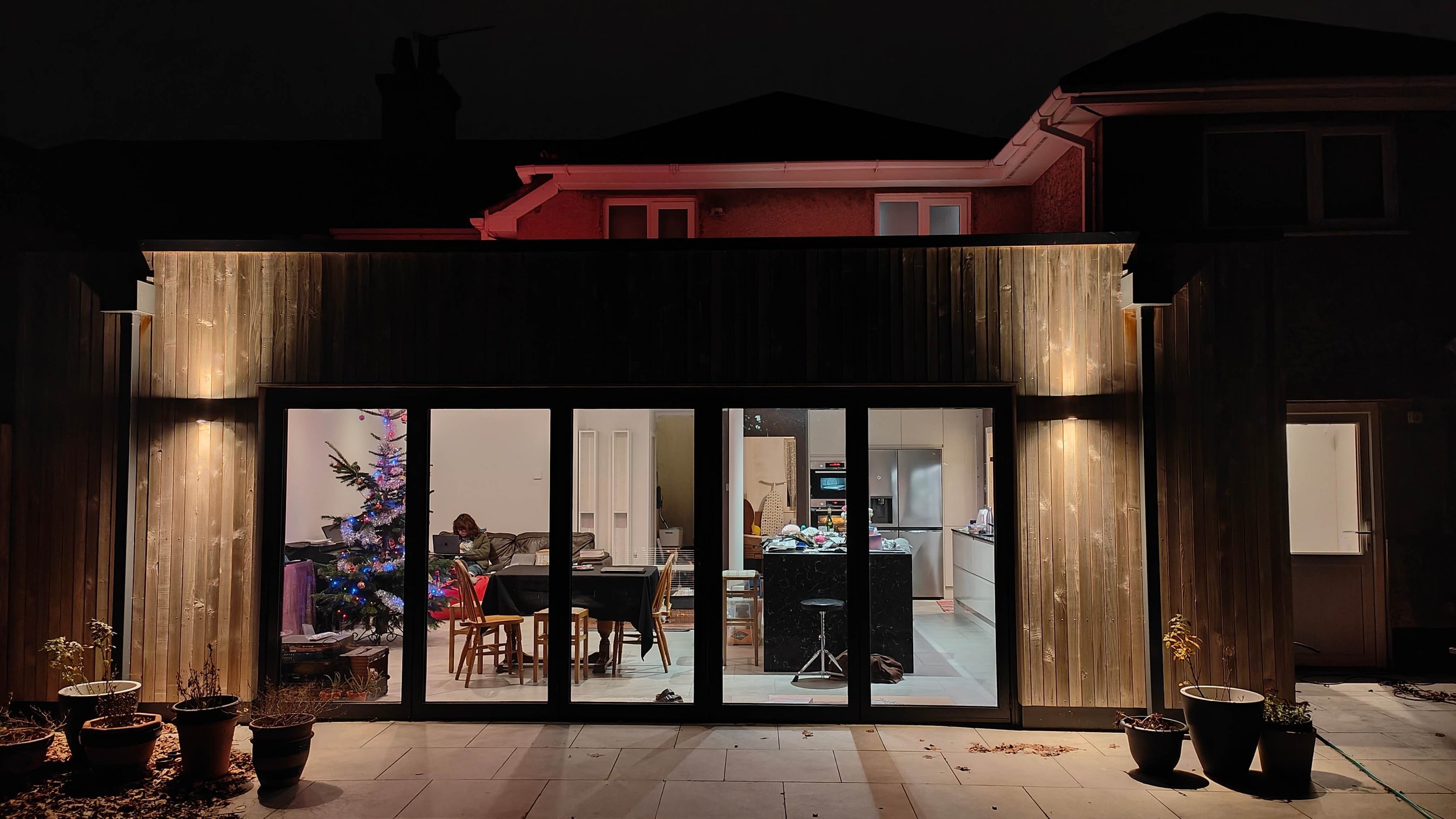 Exterior view of a modern open plan family room extension with bi-fold doors, timber cladding, kitchen island, large dining table and seating area.