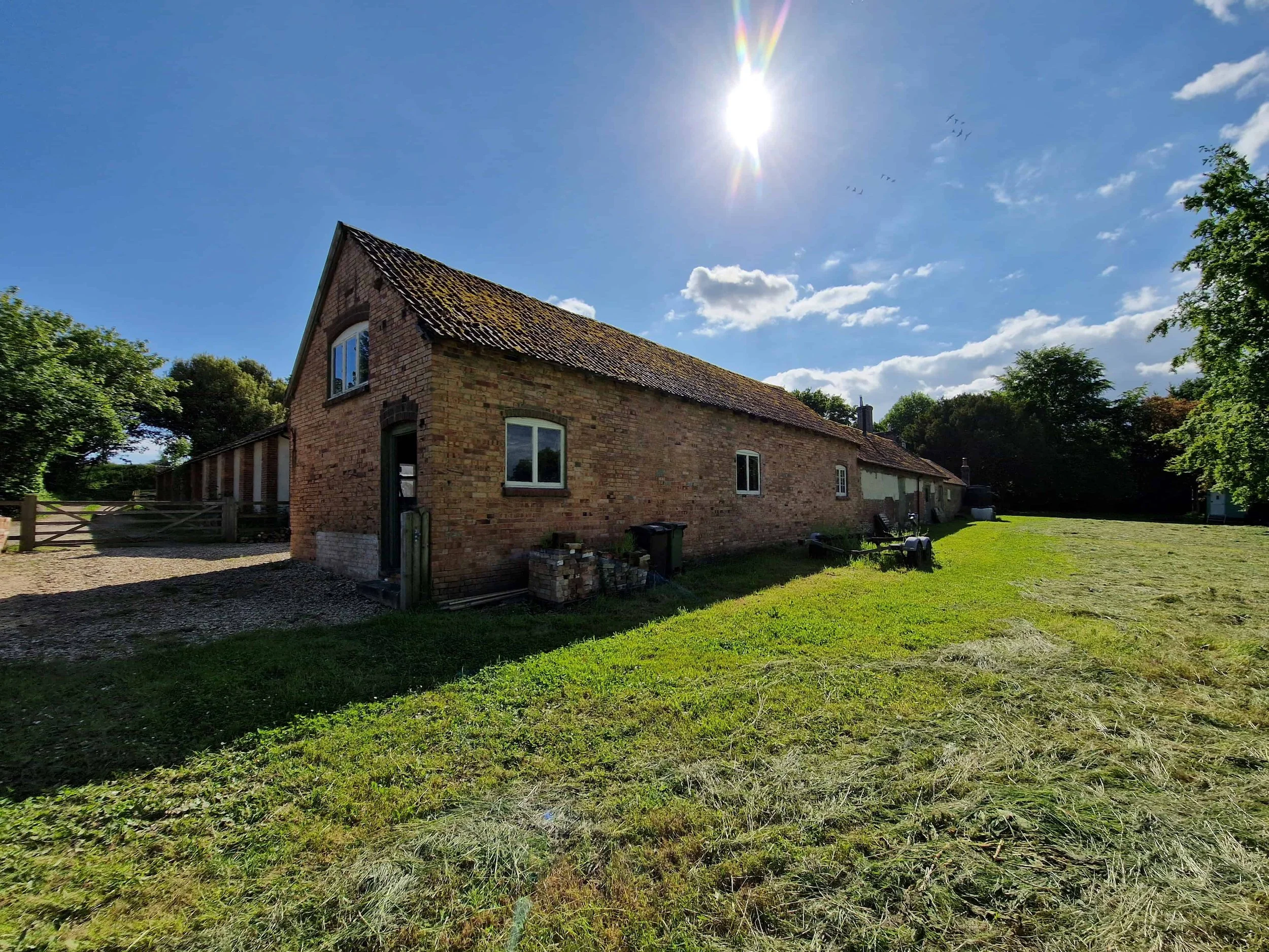 A brick walled barn in rural countryside with a pitched roof, surrounded by green trees and a grassy yard, under a bright sun in a blue sky with some clouds and birds.