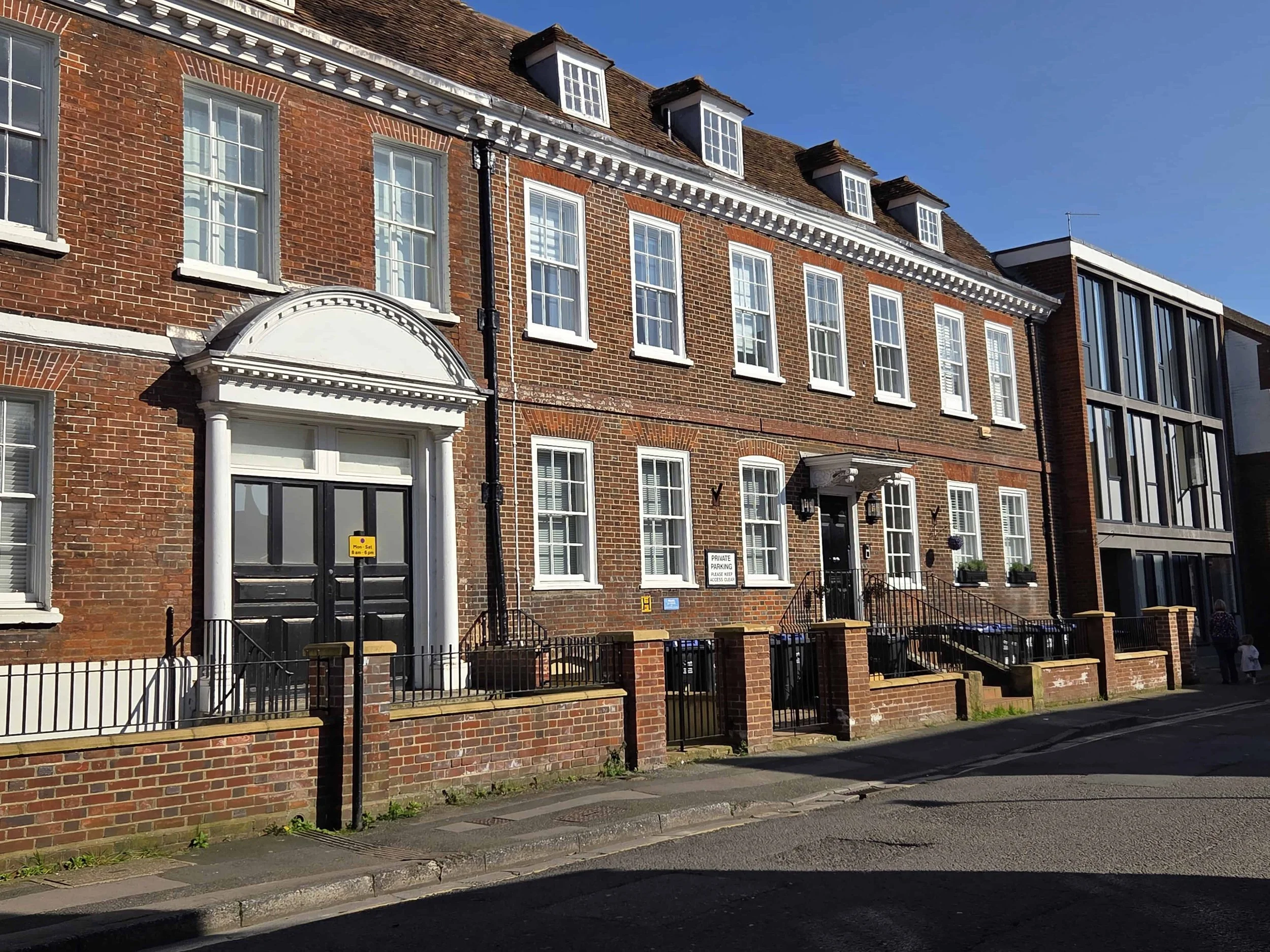 A terrace of historic traditional listed brick townhouses with sash windows and timber doors, featuring a small front yard area with black metal railings, set against a clear blue sky.