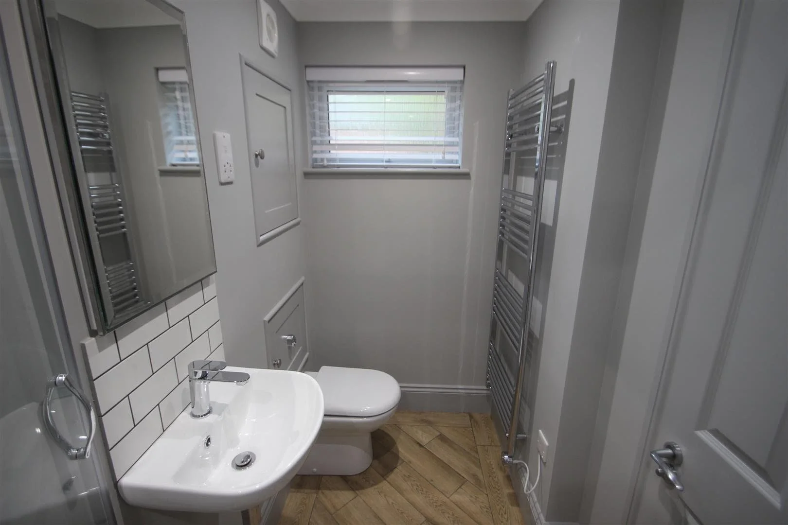 Small bathroom renovation in rental property with new wall insulation, a wall-mounted sink, a toilet, a mirror, concealed services, grey walls, a small window with blinds, and a chrome towel radiator.