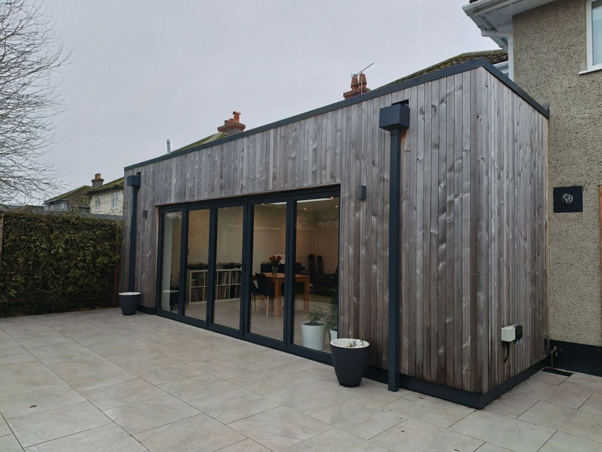 A modern rear extension on a traditional house with timber cladding and large glass doors opening to a tiled patio, with potted plants near the entrance.