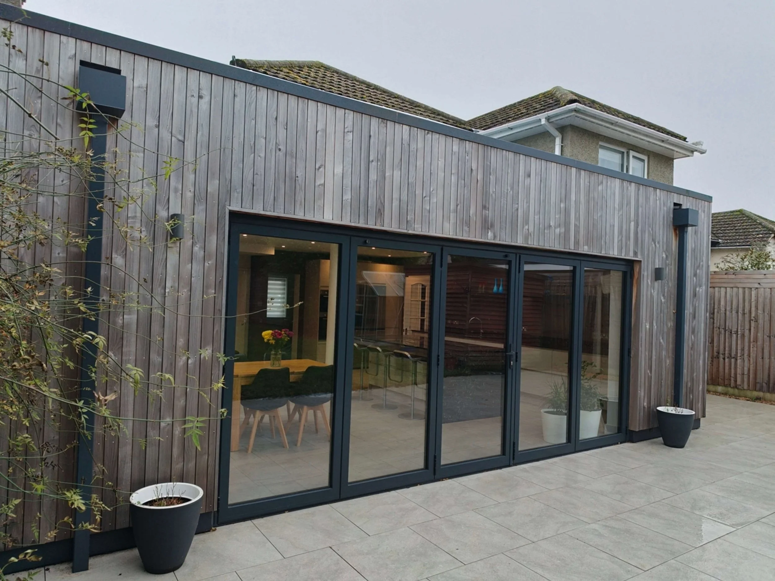 Contemporary flat roof extension with parapet roof, vertical timber cladding to exterior walls, large frameless rooflights, large bi-fold door and paved patio.