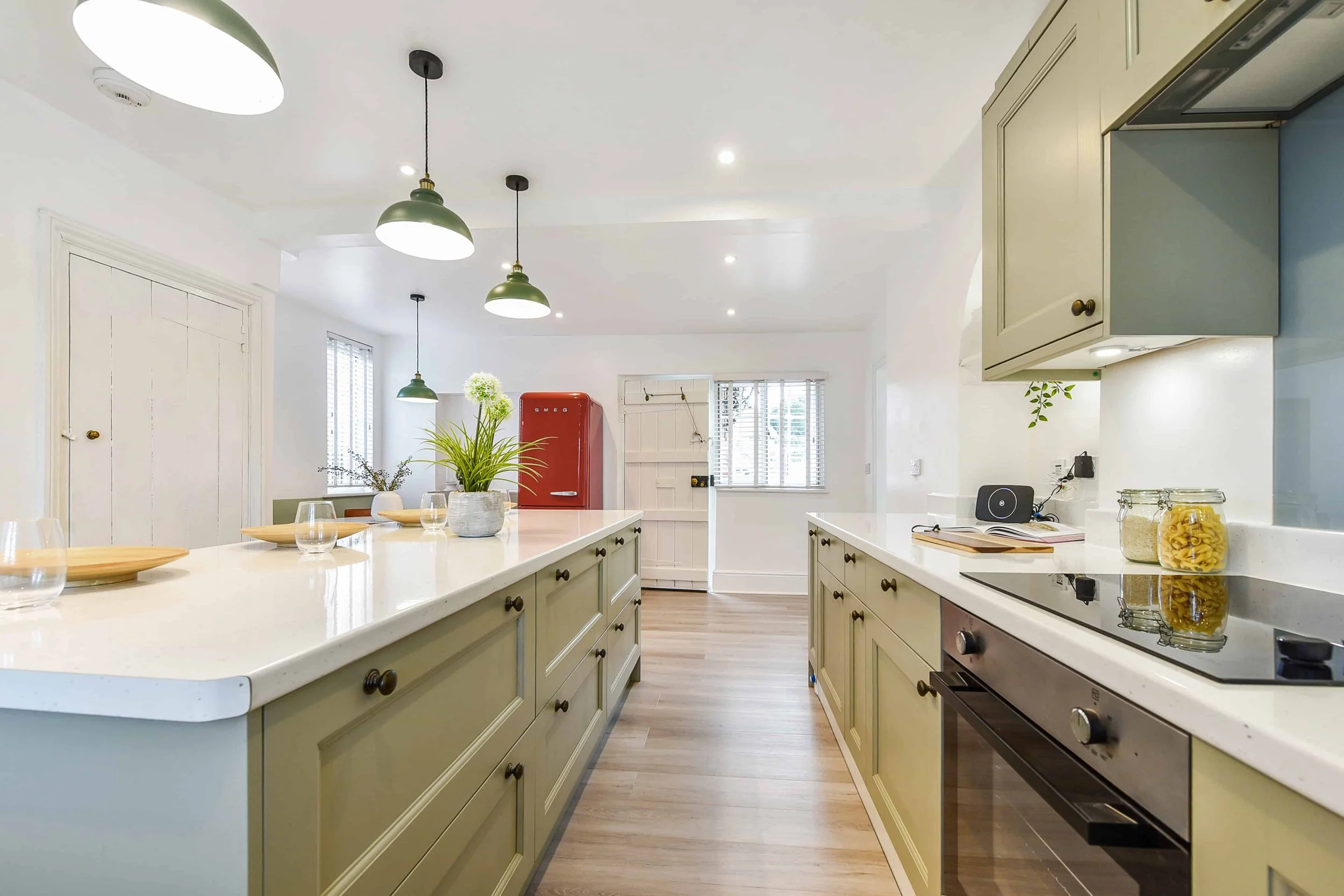 A modern galley kitchen with island, timber cabinets, marble worktops, pastel green cabinets, pendant feature lights, reclaimed timber flooring, American refrigerator, and decorative plants.