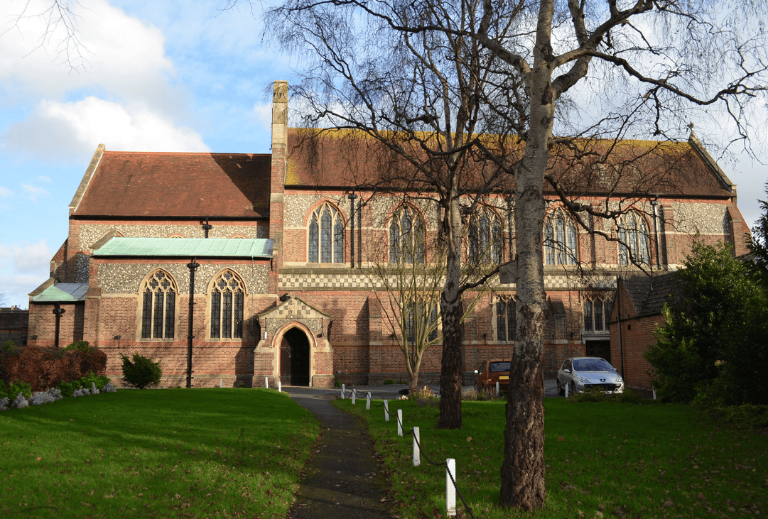 A large historic listed church with stone walls and red tiled roof, surrounded by a green lawn and leafless trees, under a partly cloudy sky.