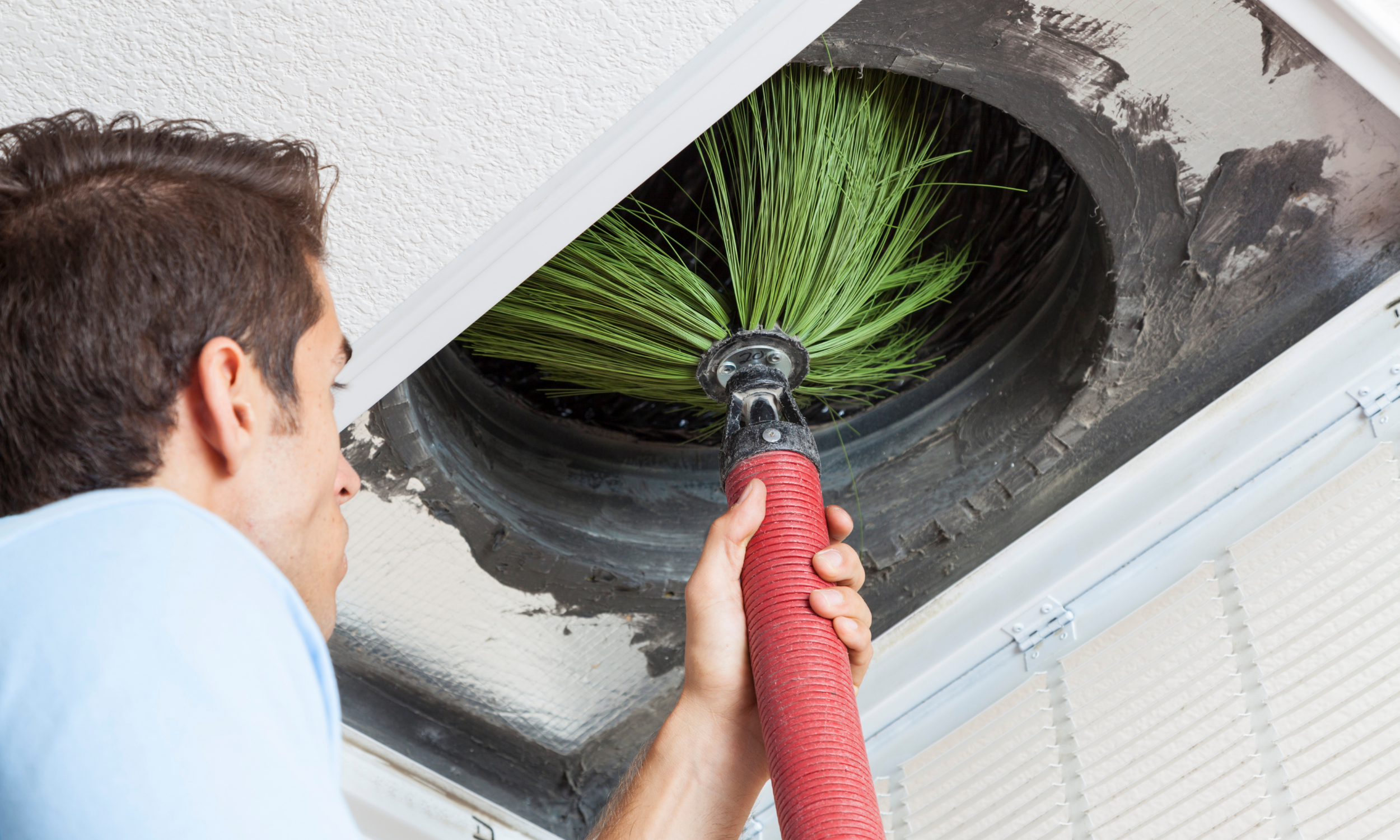 A man cleaning a ceiling vent with a green brush attachment and a red vacuum hose.