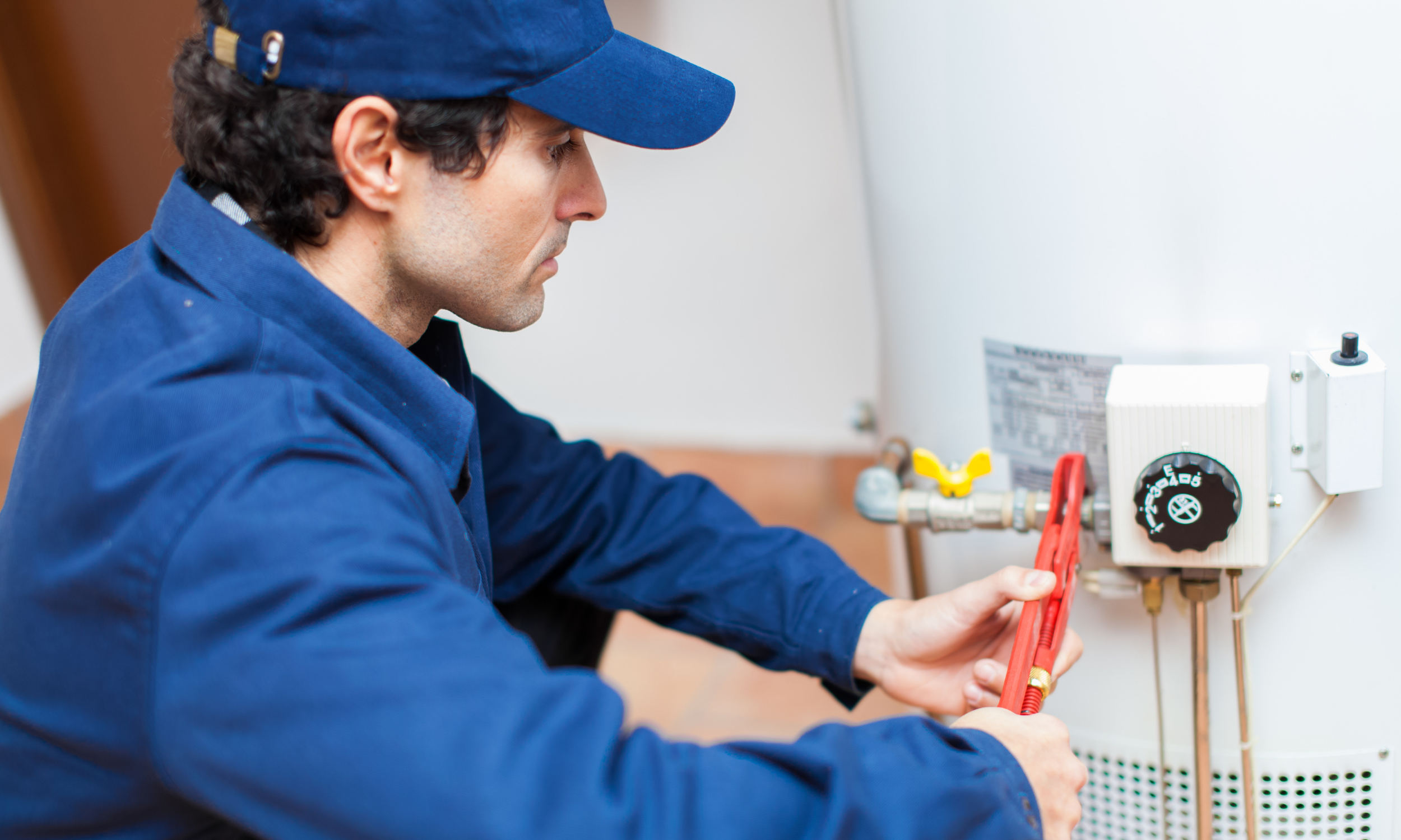 A technician in a blue uniform and cap is working on a water heater, using a red wrench.