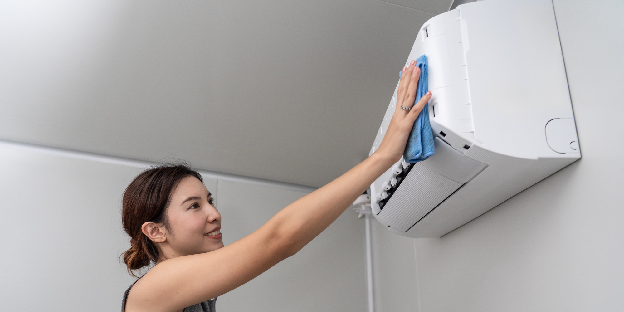 Woman cleaning the top of a wall-mounted air conditioner unit with a blue cloth.