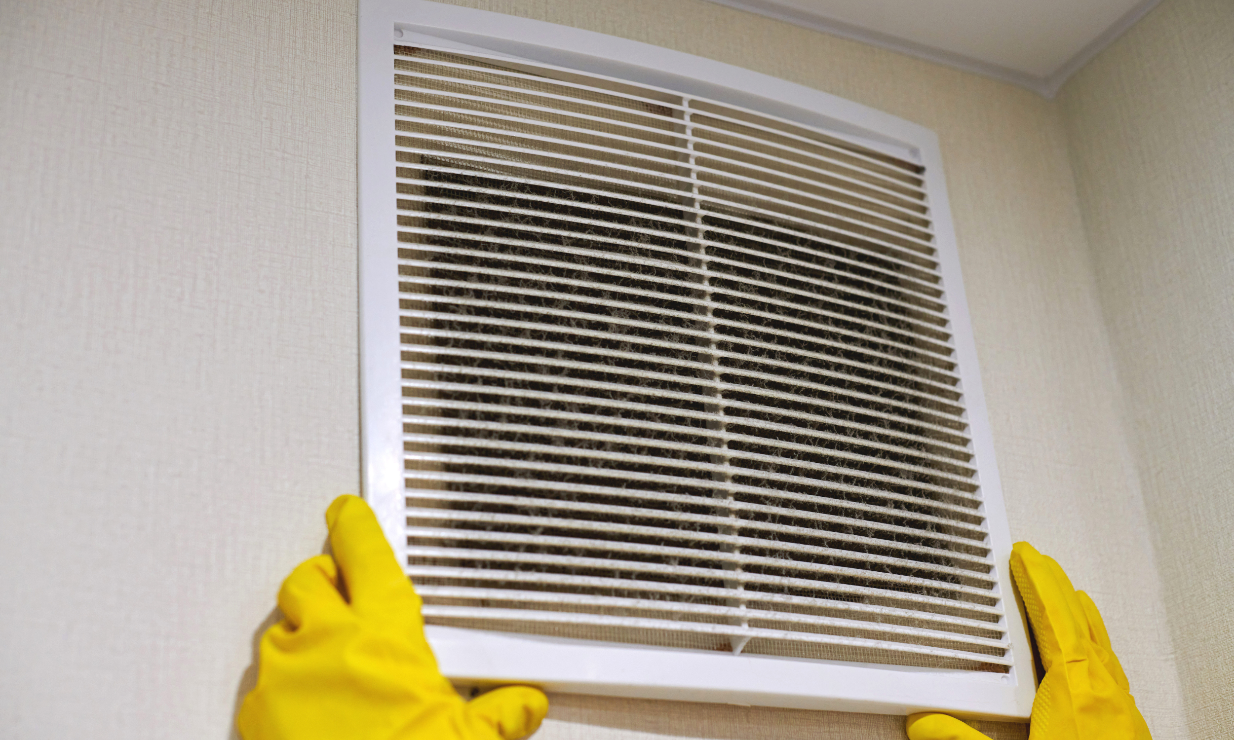 Person wearing yellow gloves removing a dirty air vent cover from a wall.