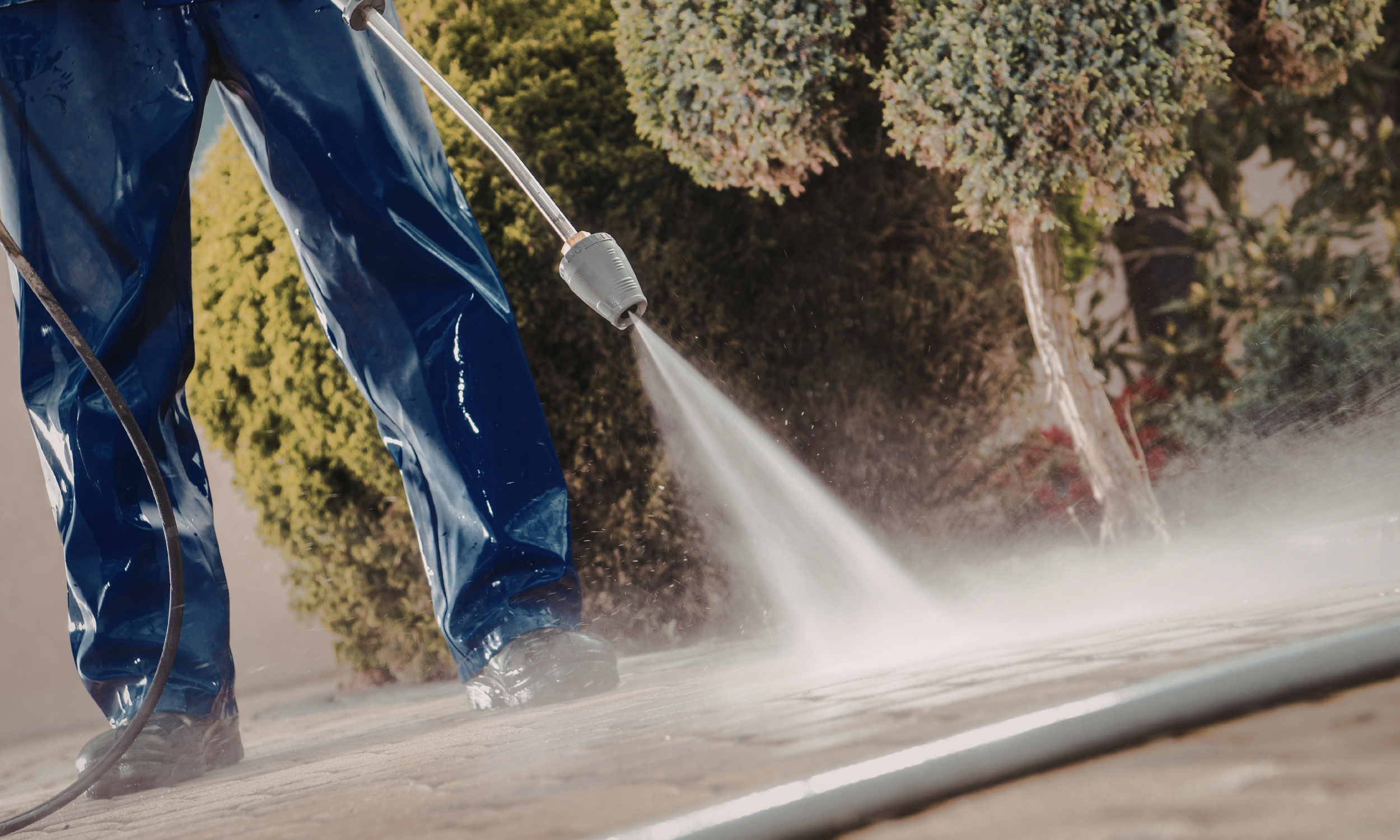 Person in blue waterproof pants using a high-pressure water spray to clean a concrete surface outdoors.