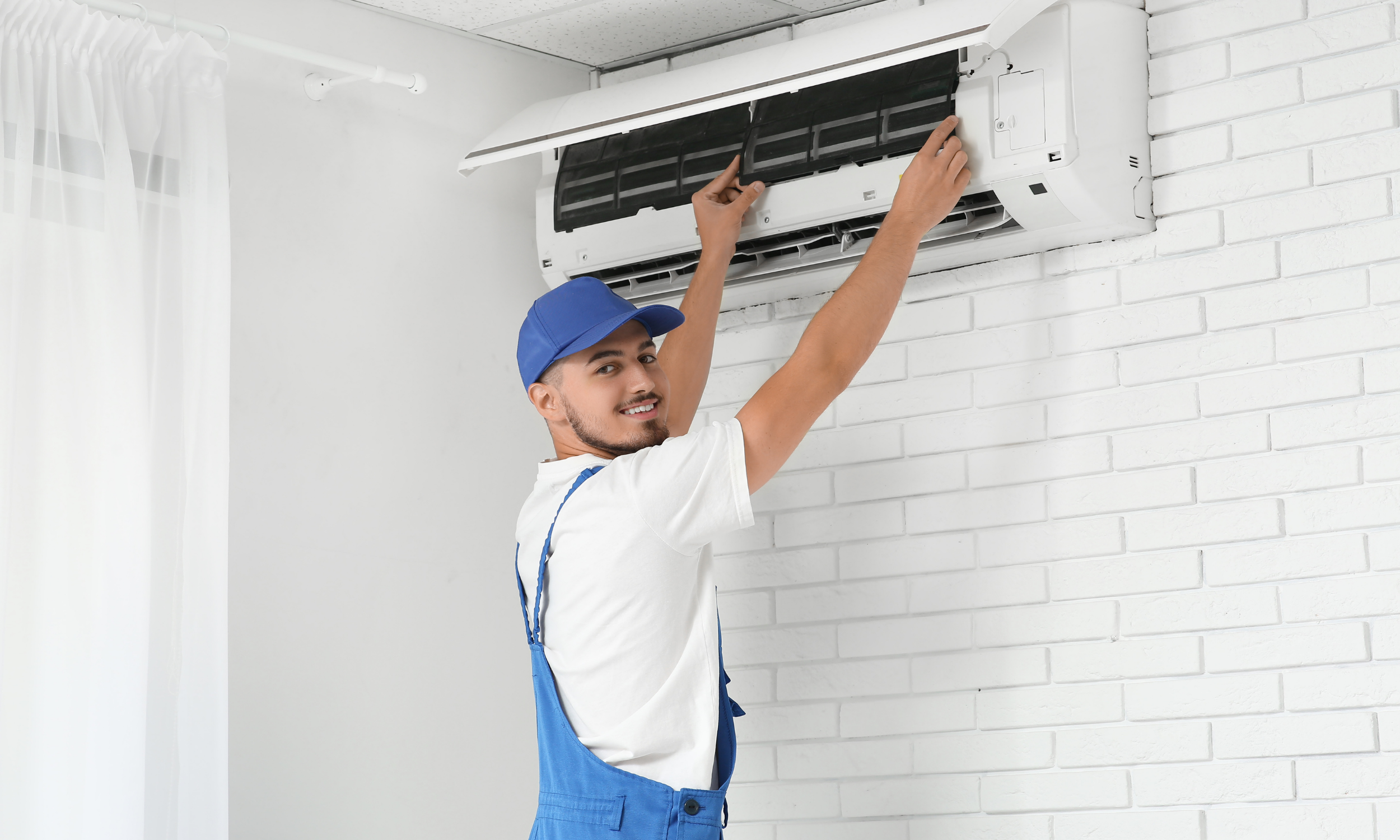 A technician in a white t-shirt and blue overalls is repairing a wall-mounted air conditioner. The technician is standing on the left side of the image, smiling, and using his hands to work on the air conditioning unit, which is embedded in a white brick wall. A white curtain is partially visible on the left side of the image.