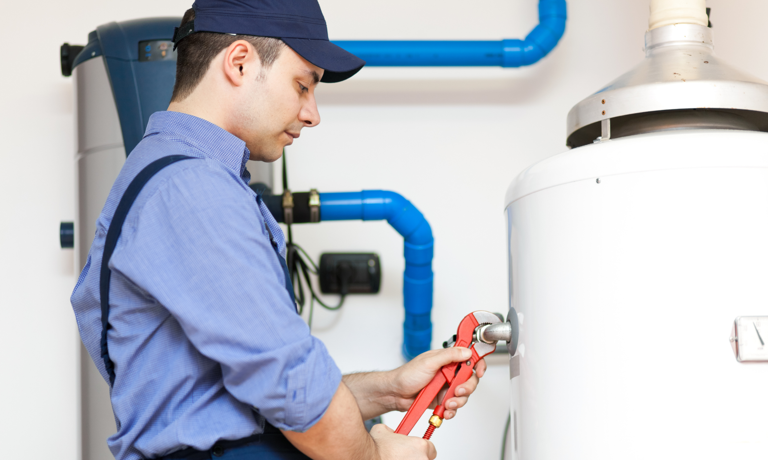 A technician in a blue shirt and dark blue baseball cap adjusts a valve on a white water heater with a red pipe wrench.