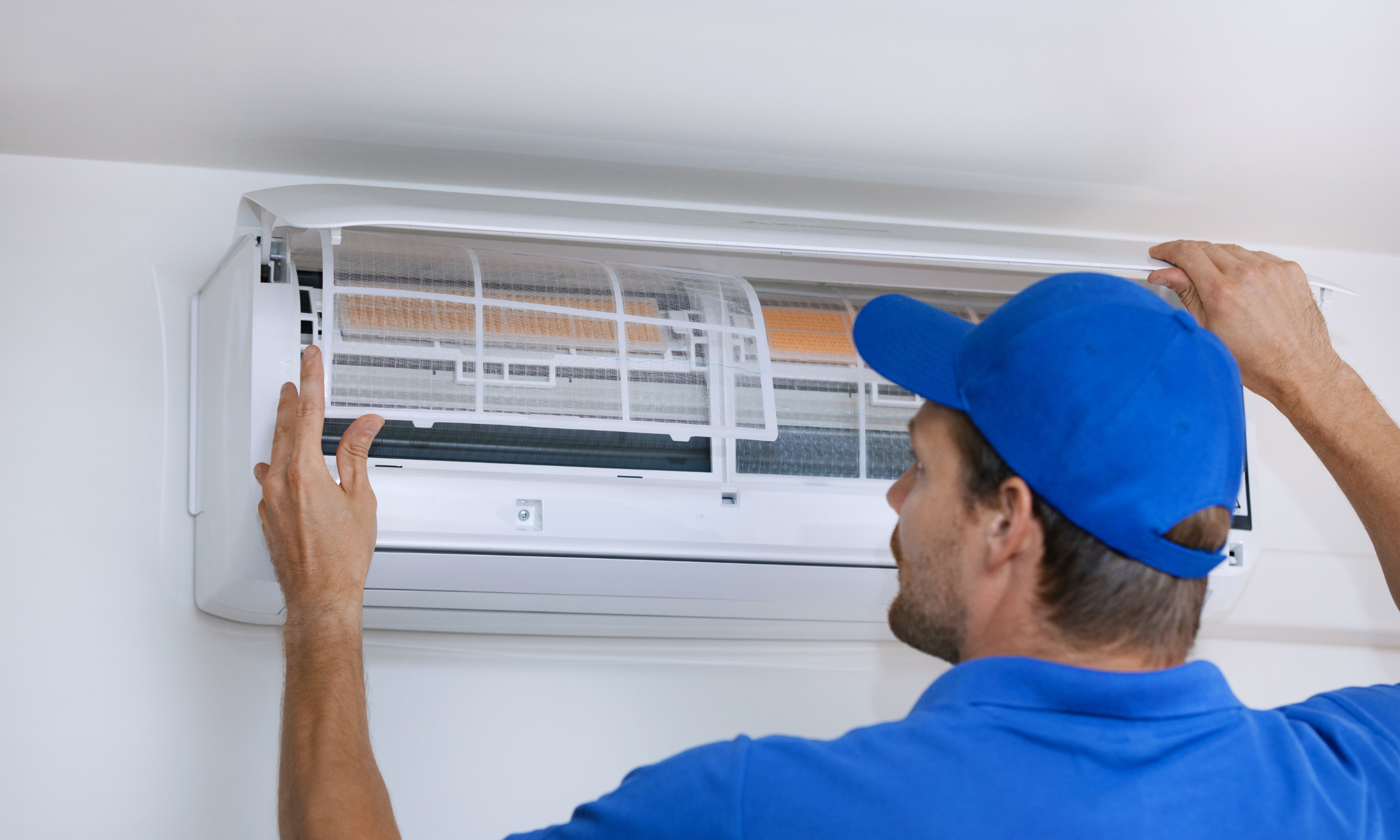 A technician in a blue cap and shirt is cleaning or repairing a wall-mounted air conditioning unit.