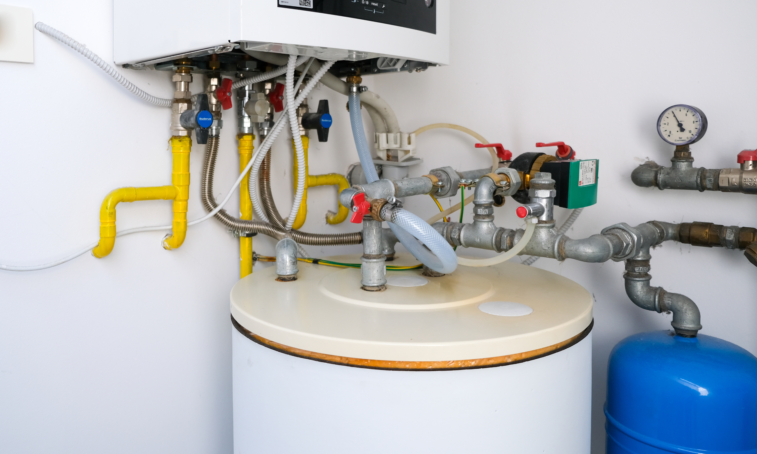 Close-up of a heating system with pipes, valves, gauges, and a storage tank installed on a white wall.