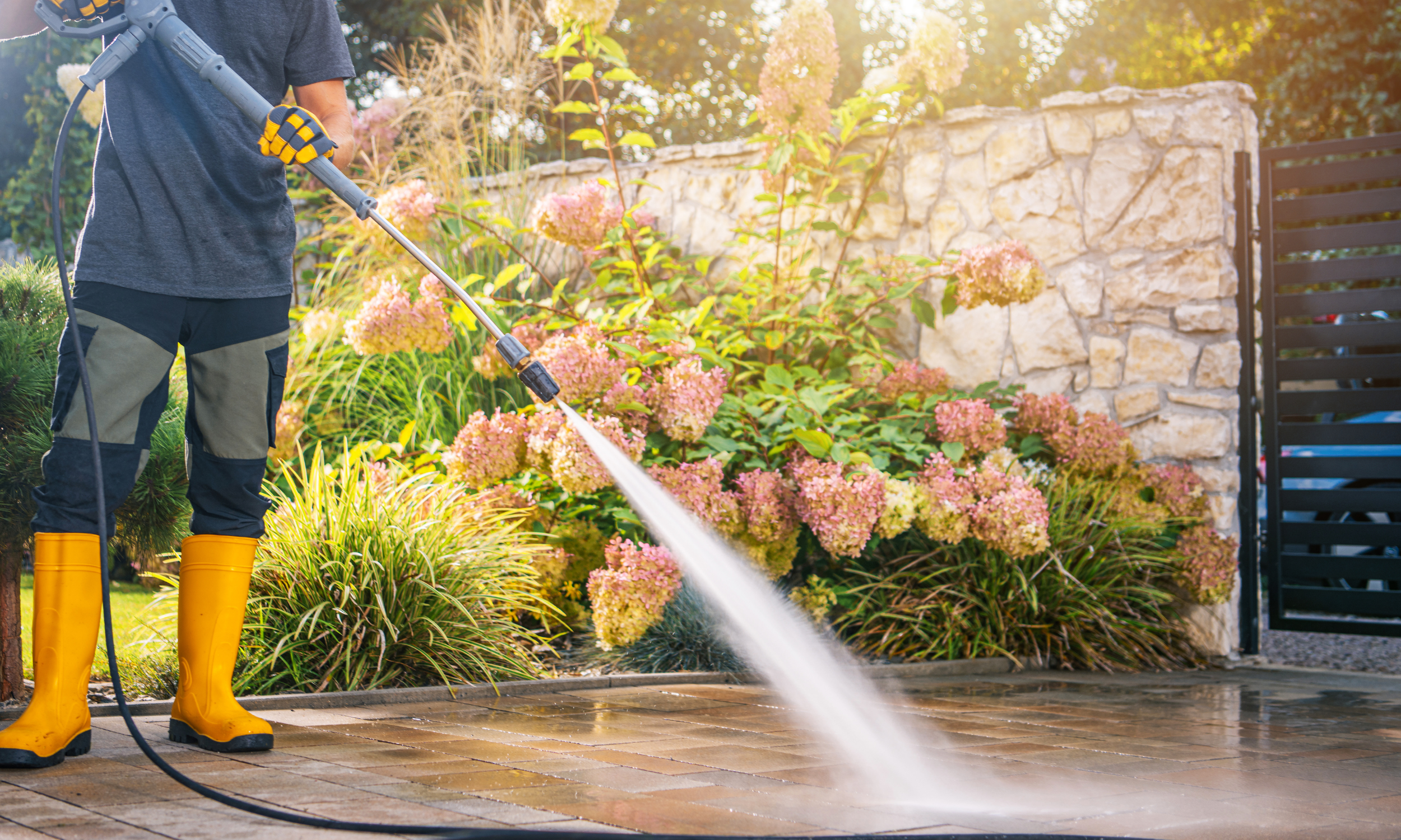 Person wearing yellow rubber boots is pressure washing a stone patio in a garden with flowering plants and bushes.