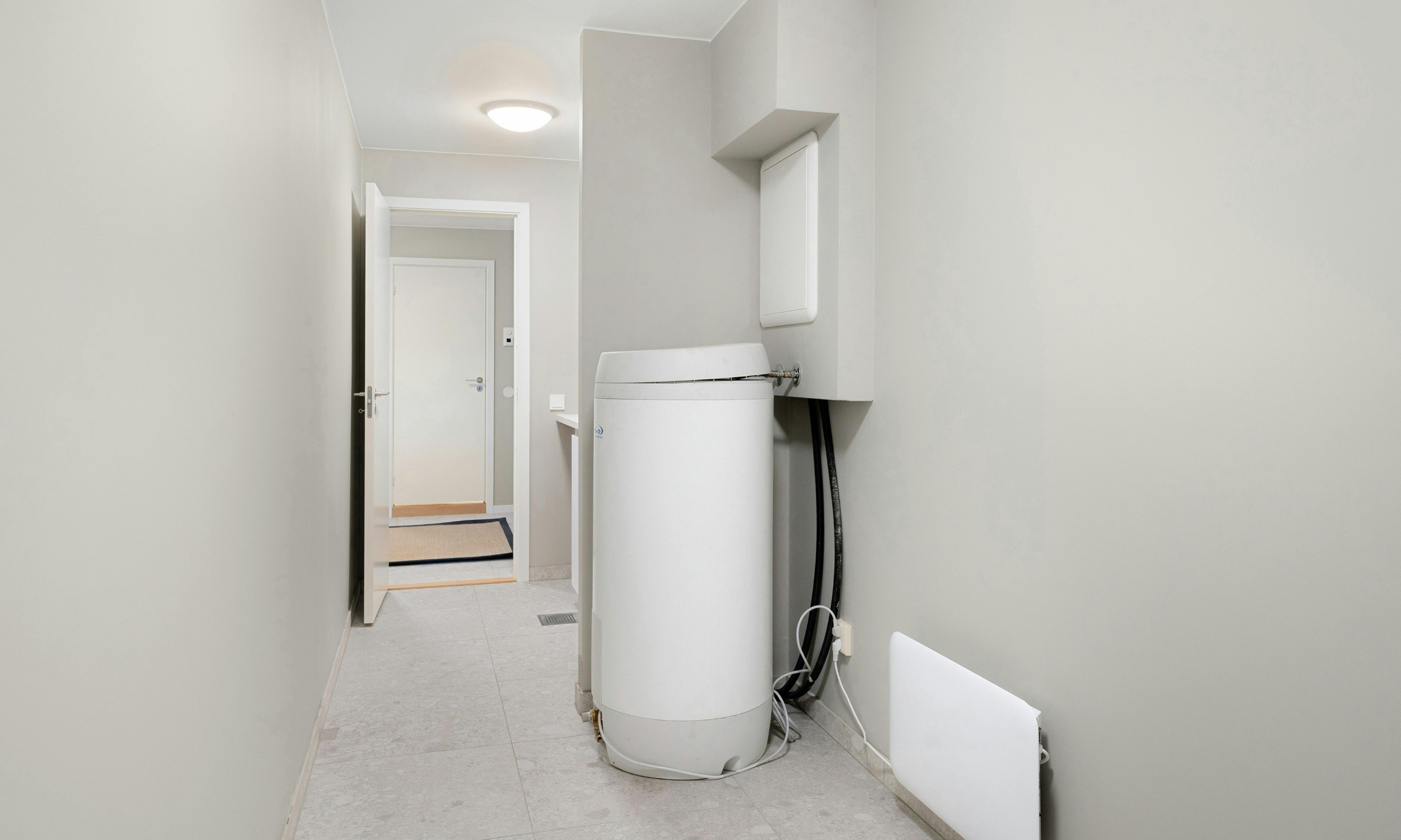 A laundry area with a white top-loading washing machine, electrical cords, and a wall-mounted cabinet inside a room with light-colored walls and tiled flooring.