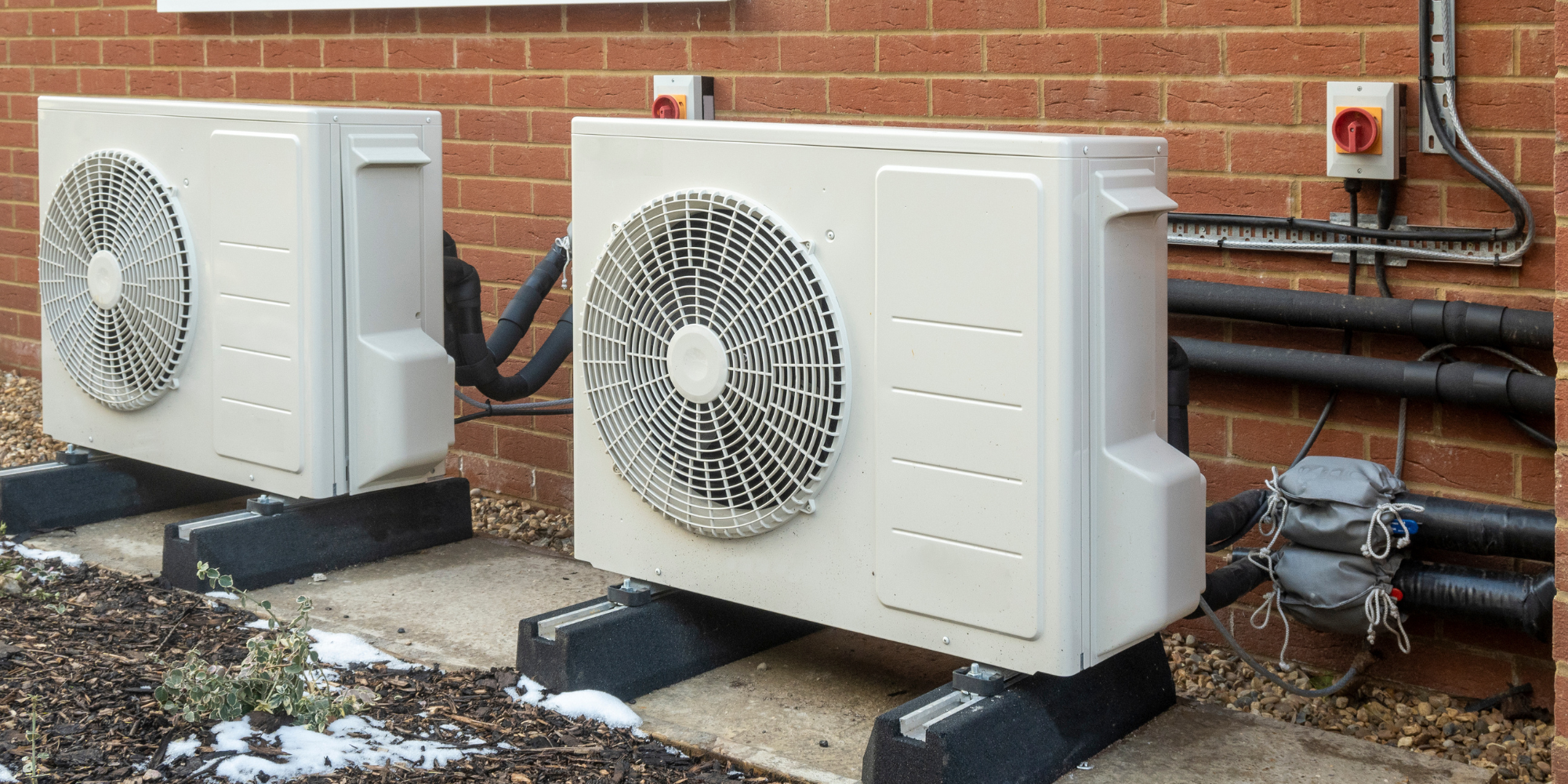 Two white outdoor HVAC units installed on a concrete pad outside a brick building, with black pipes and electrical wiring connected to them.