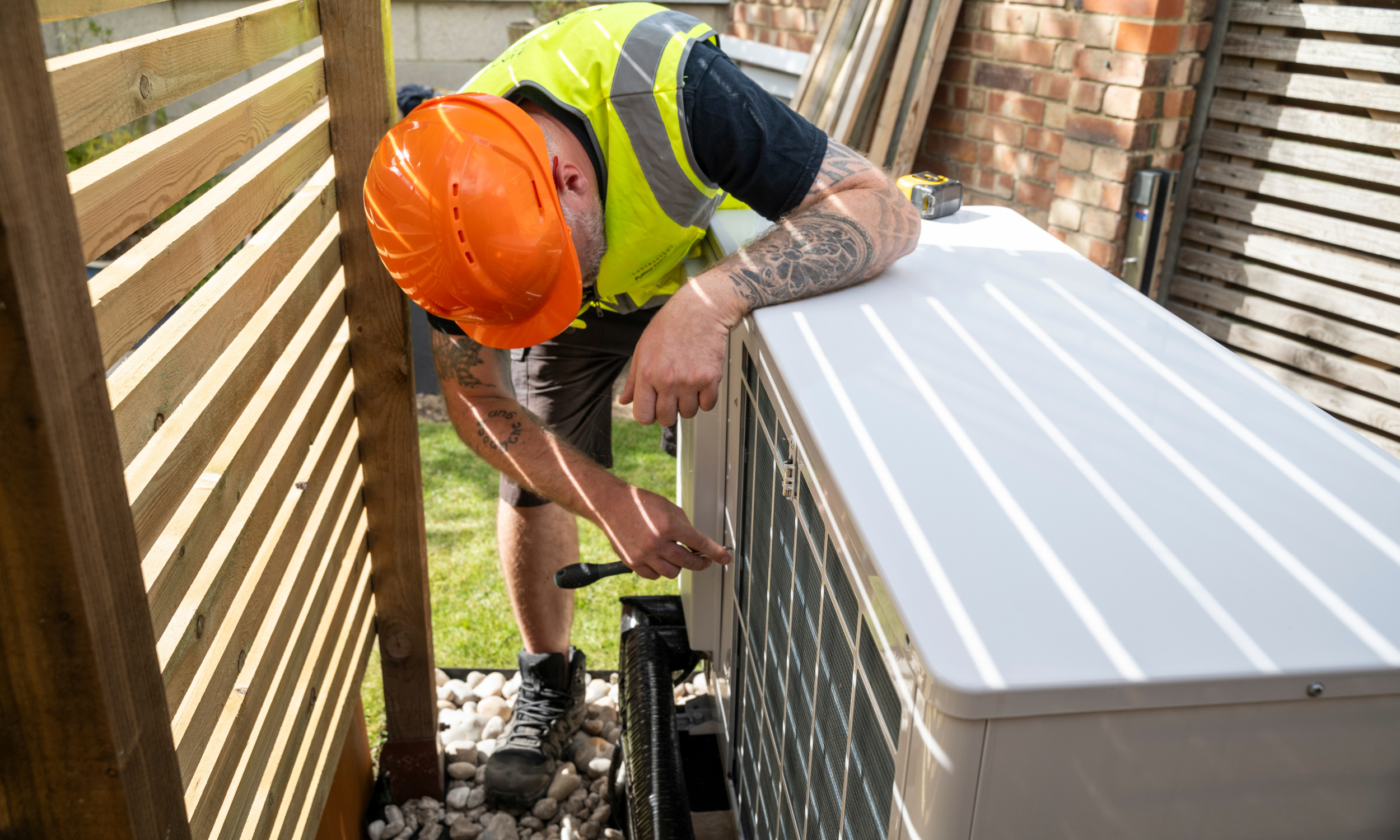 A worker wearing an orange safety helmet, a neon yellow safety vest, and black work boots is repairing or installing an air conditioning unit outdoors.