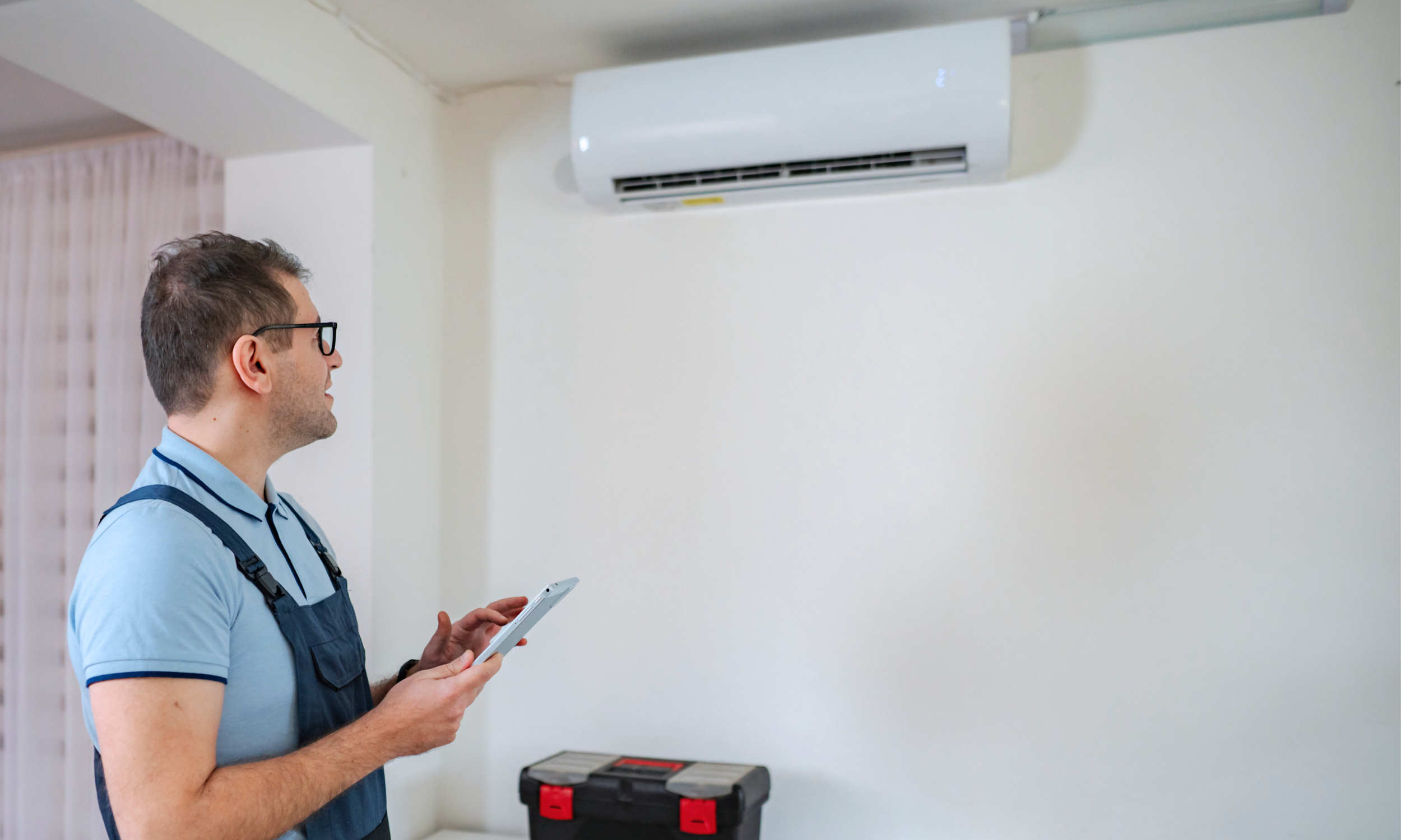 A man with glasses, wearing a blue shirt and work vest, holding a tablet, standing in a room with a wall-mounted air conditioner.