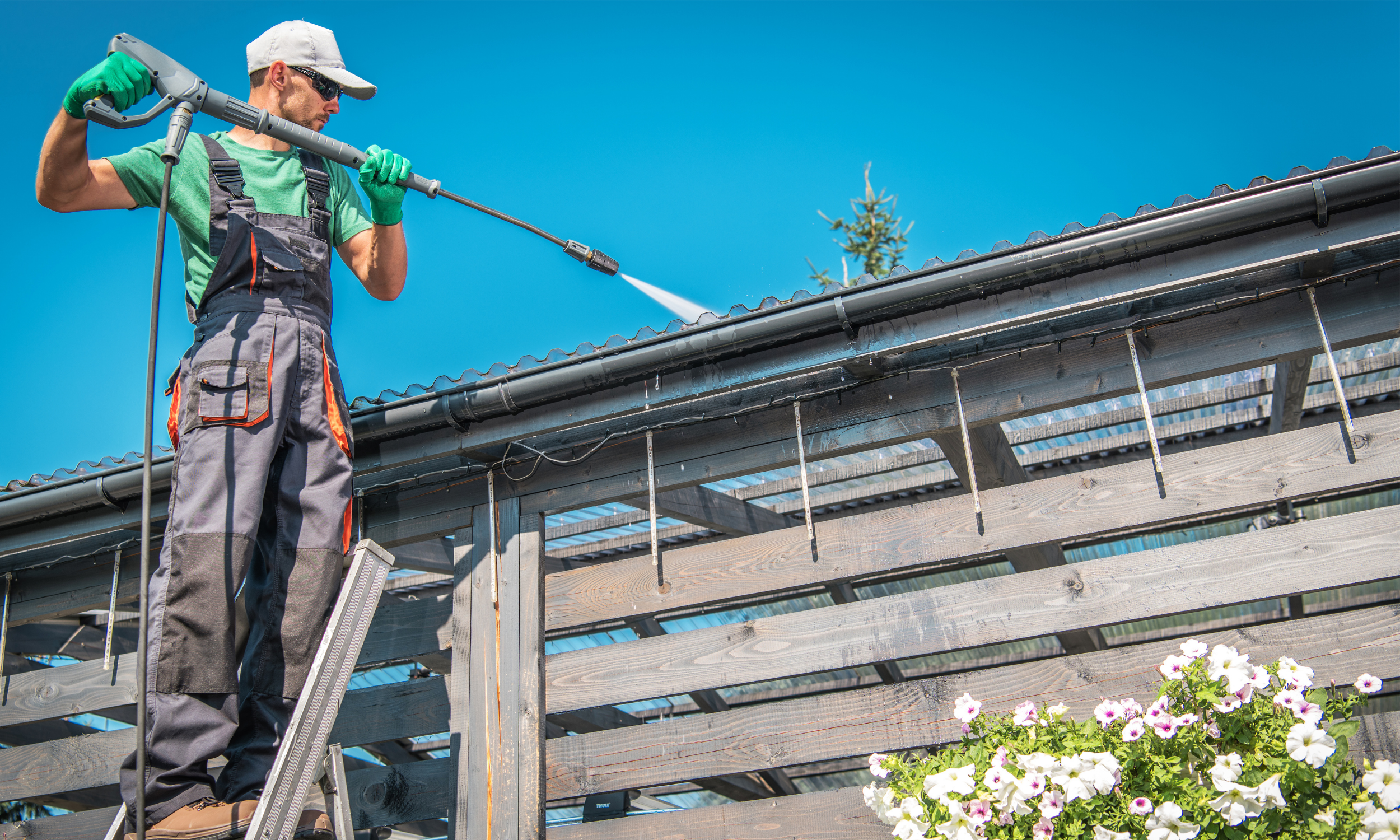 A man wearing a gray cap, sunglasses, green t-shirt, and work overalls is pressure washing a wooden structure outdoors under a clear blue sky.
