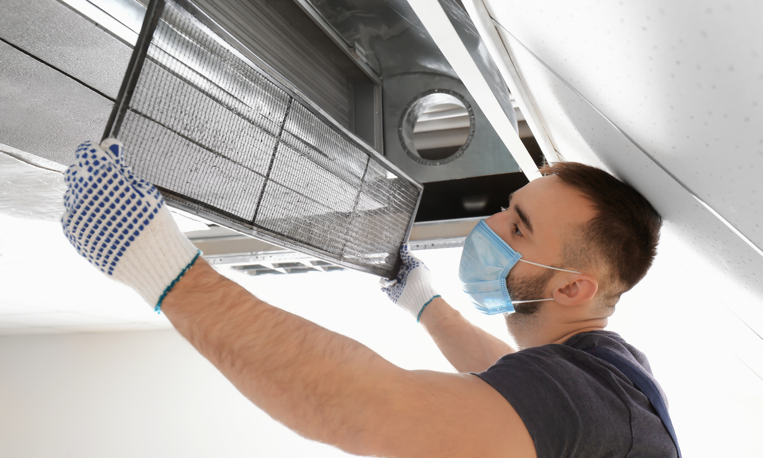 A man wearing a face mask and gloves is installing or inspecting an air vent or exhaust fan on the ceiling.