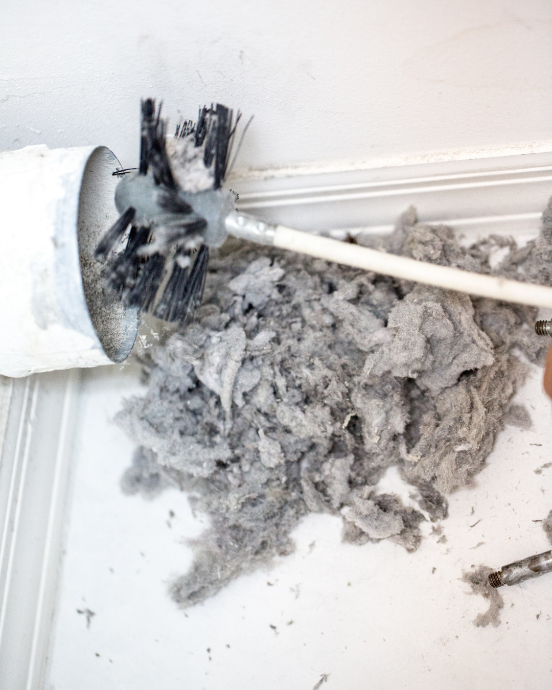 A vent cleaning brush cleaning dust and debris from a vent in the wall of a room with drywall and molding.