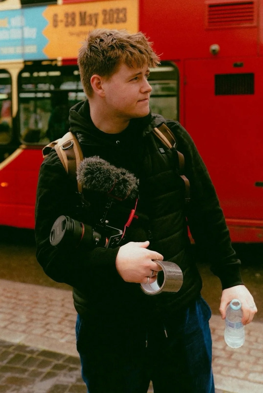 Young man holding a video camera, water bottle, and tape, standing in front of a red bus.