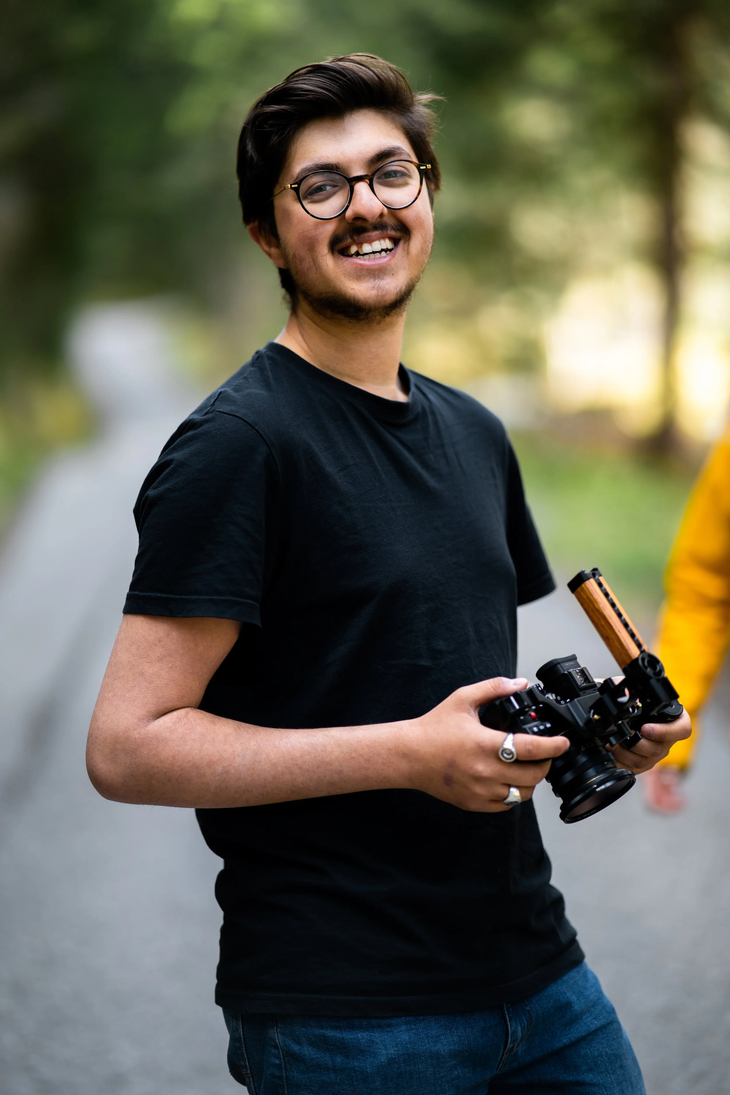 A young man with glasses and a beard, smiling, holding a camera outdoors on a path in a forested area.
