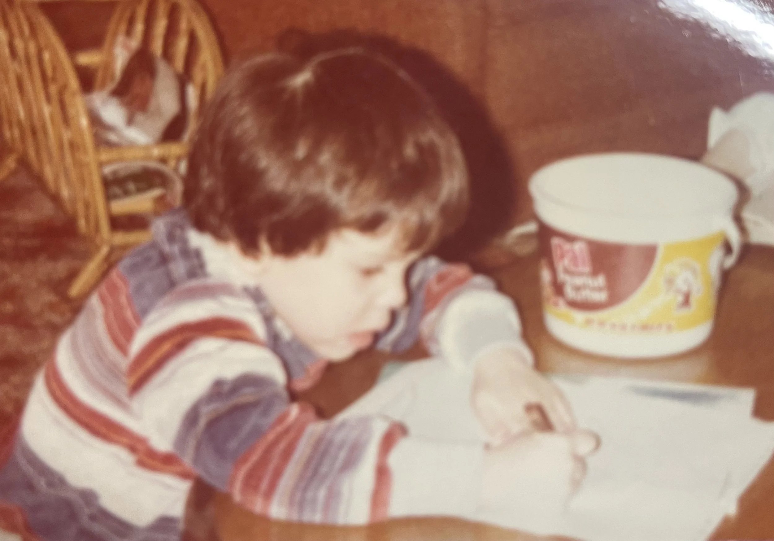A small child with brown hair, wearing a striped shirt, is reaching for a container of Yoplait yogurt on a table. There is a woven basket nearby, and the child appears to be in a kitchen or dining area.