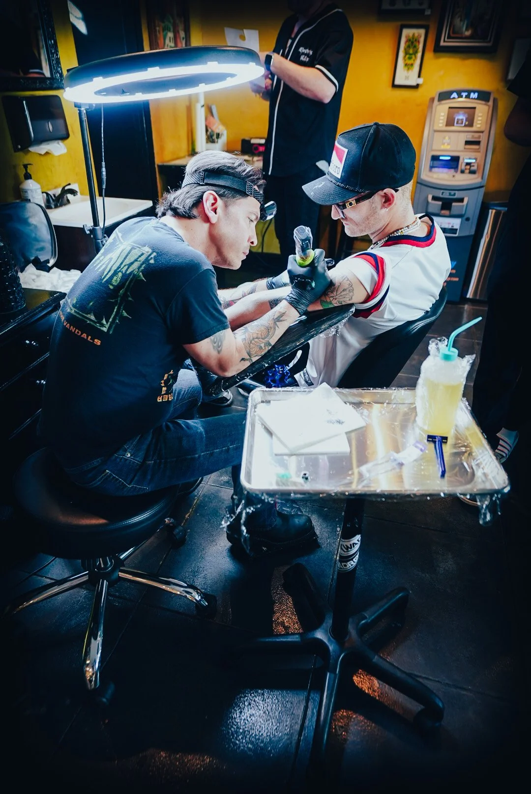 A person getting a tattoo on their arm in a tattoo studio, with an artist working on their tattoo under a bright light. The artist has tattoos on his arms and wears a black T-shirt. The client is wearing a cap and glasses, and there are supplies on a tray nearby.