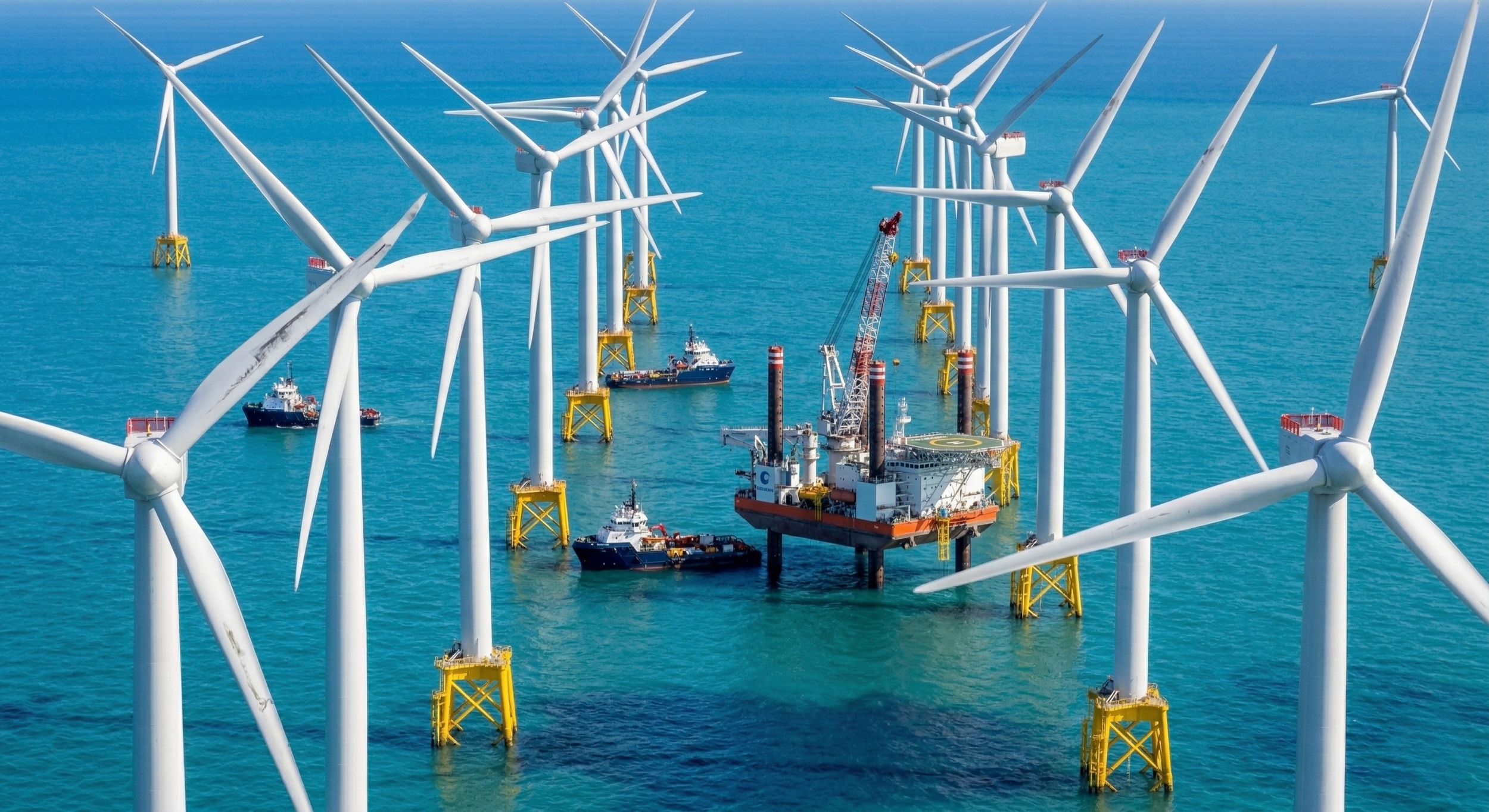 Offshore wind farm with multiple wind turbines in the ocean, supported by yellow structures, with ships and a platform in the water.