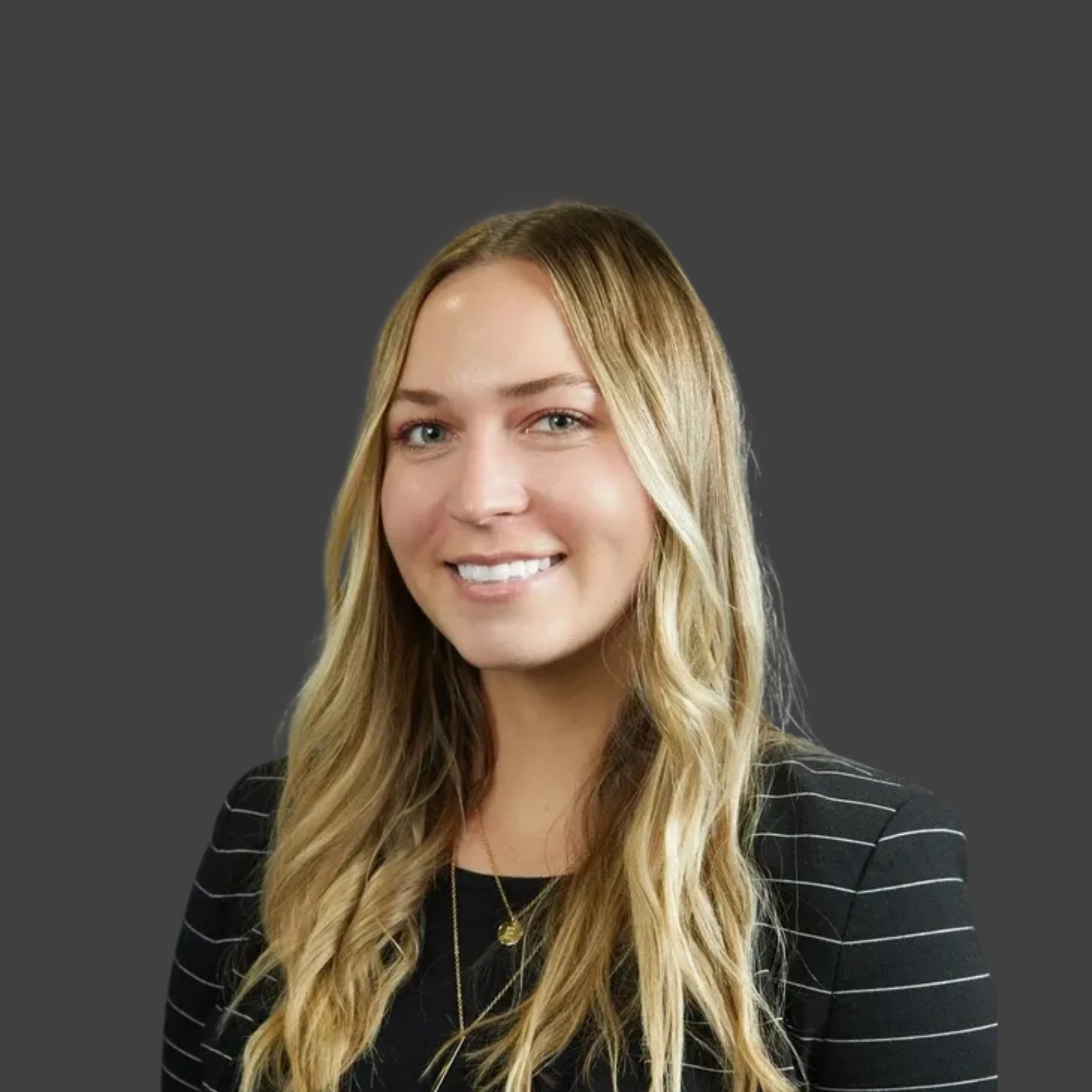 Professional headshot of a smiling woman with long, wavy blonde hair, wearing a black blazer with white pinstripes and a gold necklace, against a gray background.