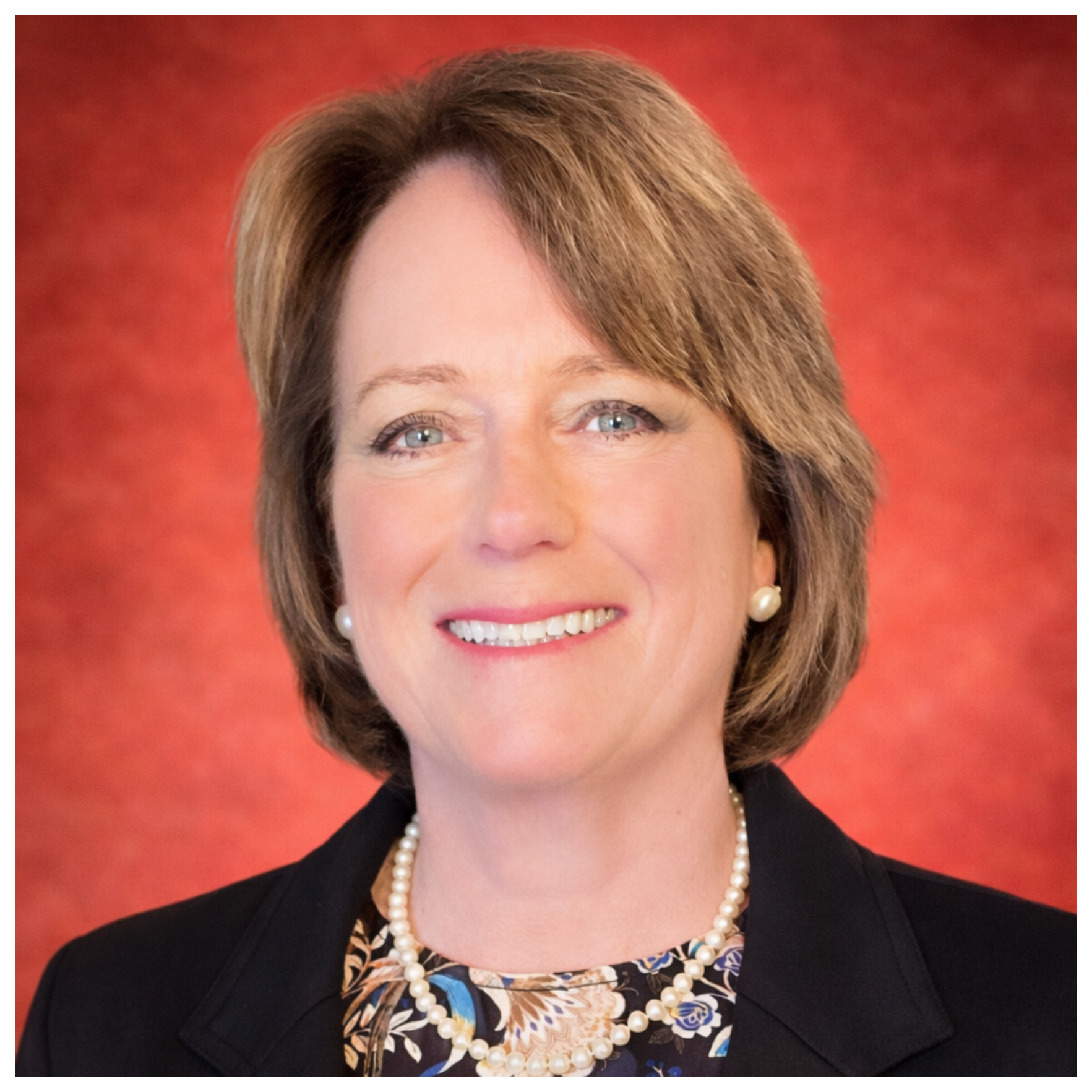A professional woman with short brown hair, wearing pearl earrings, a pearl necklace, and a floral blouse under a black blazer, smiling against a red background.