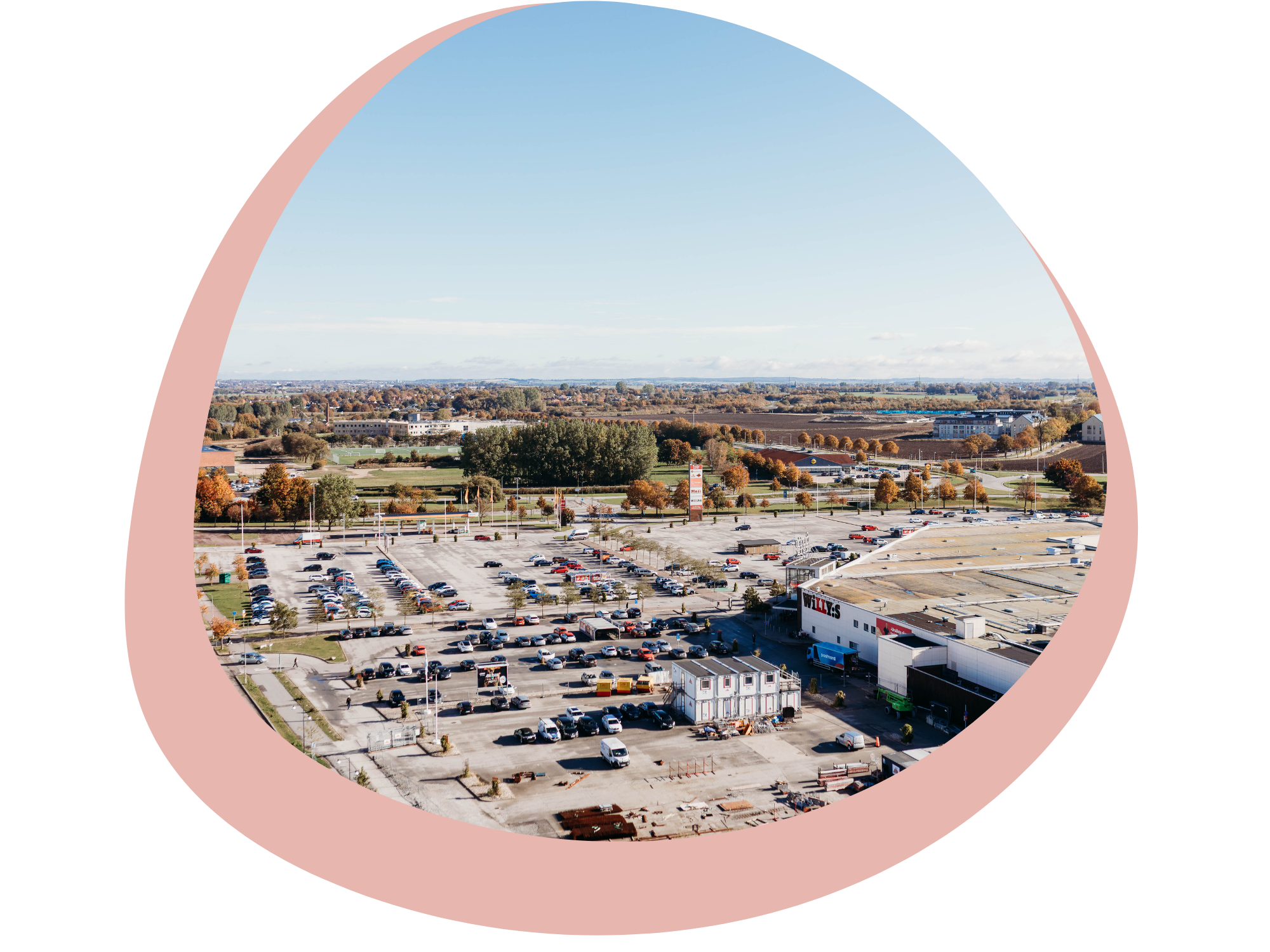 En översiktsbild av ett parkeringsområde med många bilar, ett köpcentrum och ett landskap med träd och öppna fält. Himlen är blå med några moln och flygplan kan ses i fjärran.