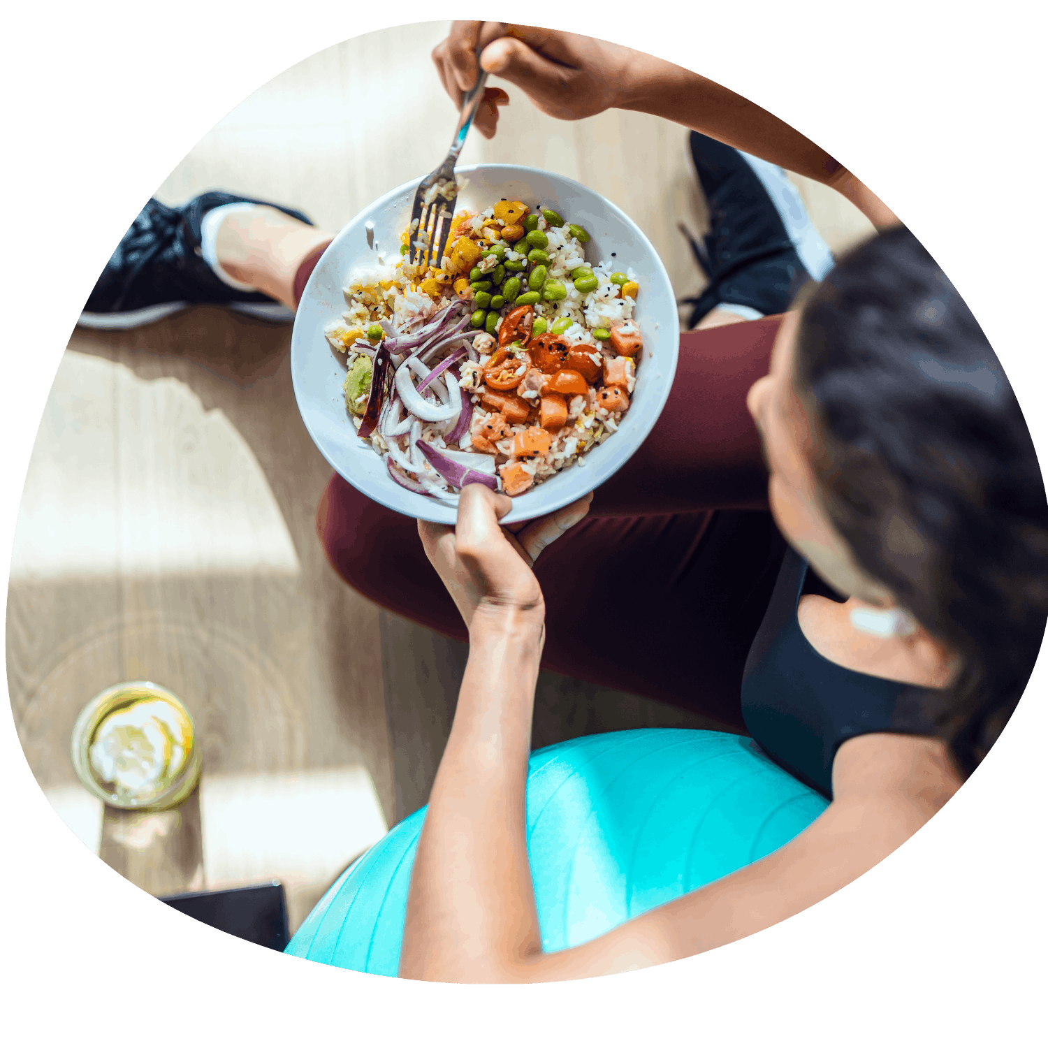 Person som håller en skål med sallad, innehållande grönsaker och ris, sittande på golvet med ett glas citronvatten bredvid. Personen är klädd i ljusblått byxor och en svart topp.