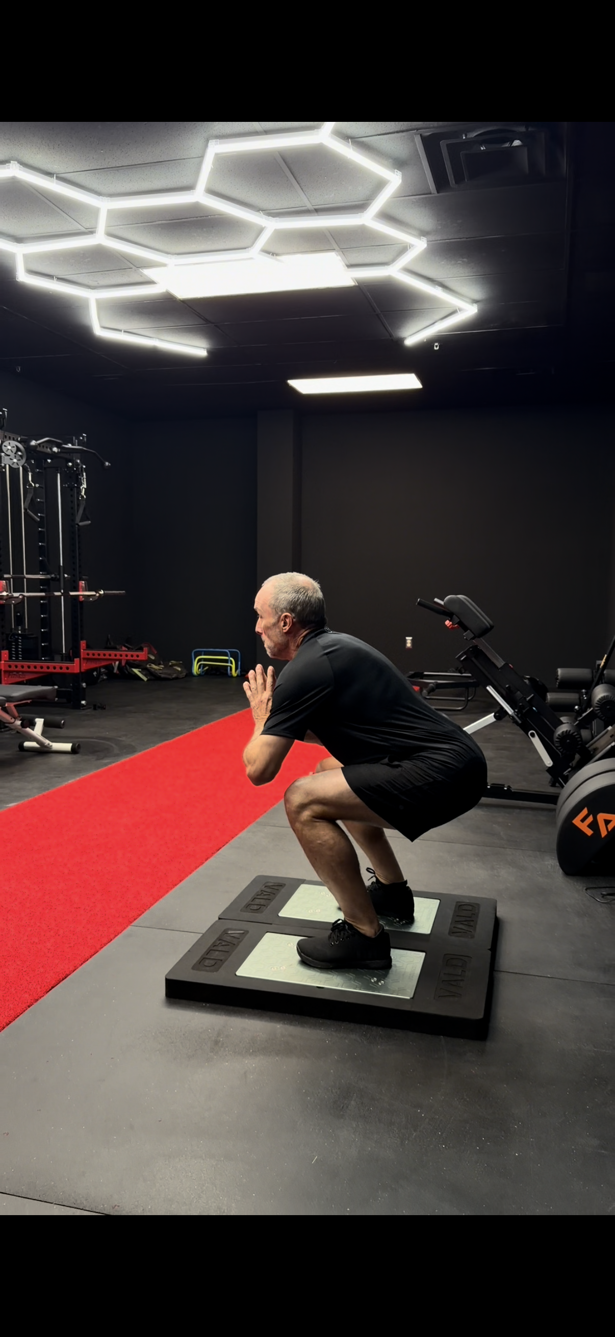 A man is squatting on a workout platform in a gym, with his hands pressed together in front of his chest, performing a fitness exercise.