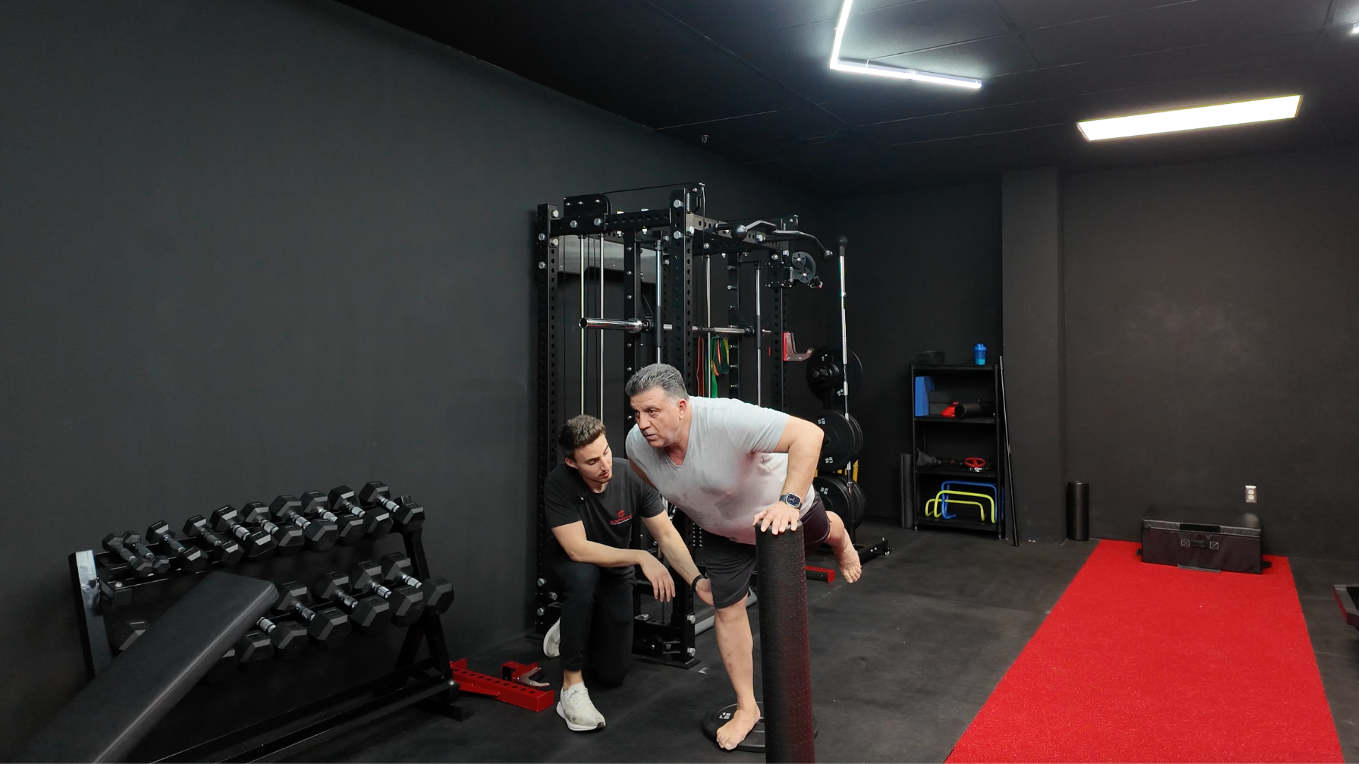 An older man with gray hair doing a balance exercise on a foam roller in a gym, assisted by a woman in black sportswear.