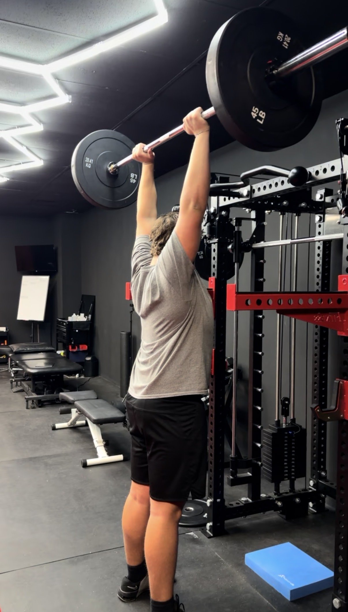 A person lifting a barbell with weights overhead in a gym, standing on a black floor with gym equipment and chairs in the background.
