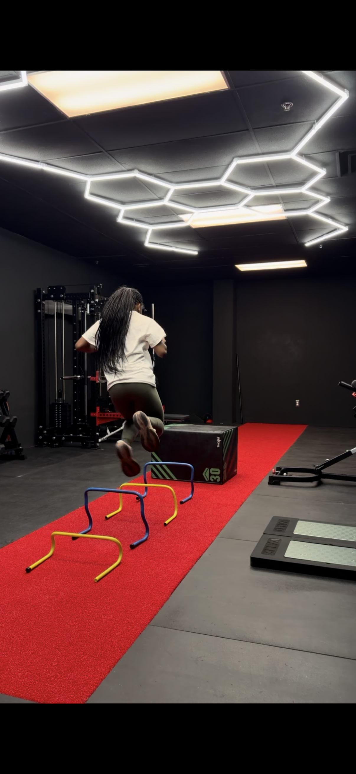 A woman is jumping over small hurdles on a red track inside a fitness gym, with exercise equipment in the background.