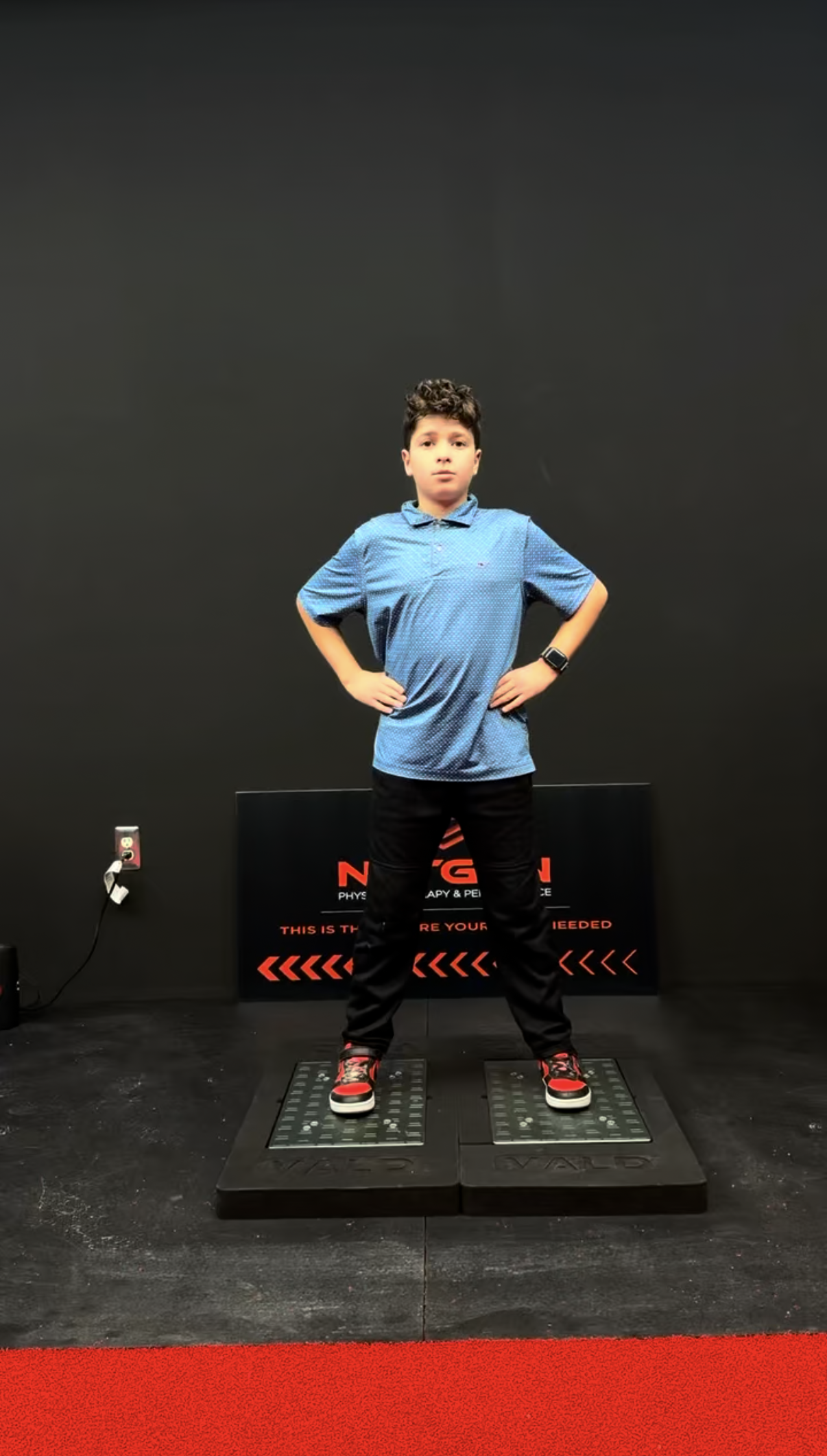 Young boy standing on a vibration plate machine in a gym or therapy center with arms akimbo, wearing a blue shirt, black pants, and colorful sneakers, with a black wall and a sign in the background.