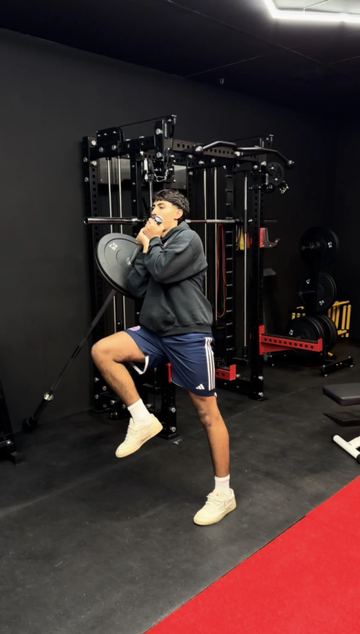 A young man in athletic shorts, a hoodie, and sneakers is exercising with a weight plate in a gym, using a landmine attachment for a lunge or squat exercise.