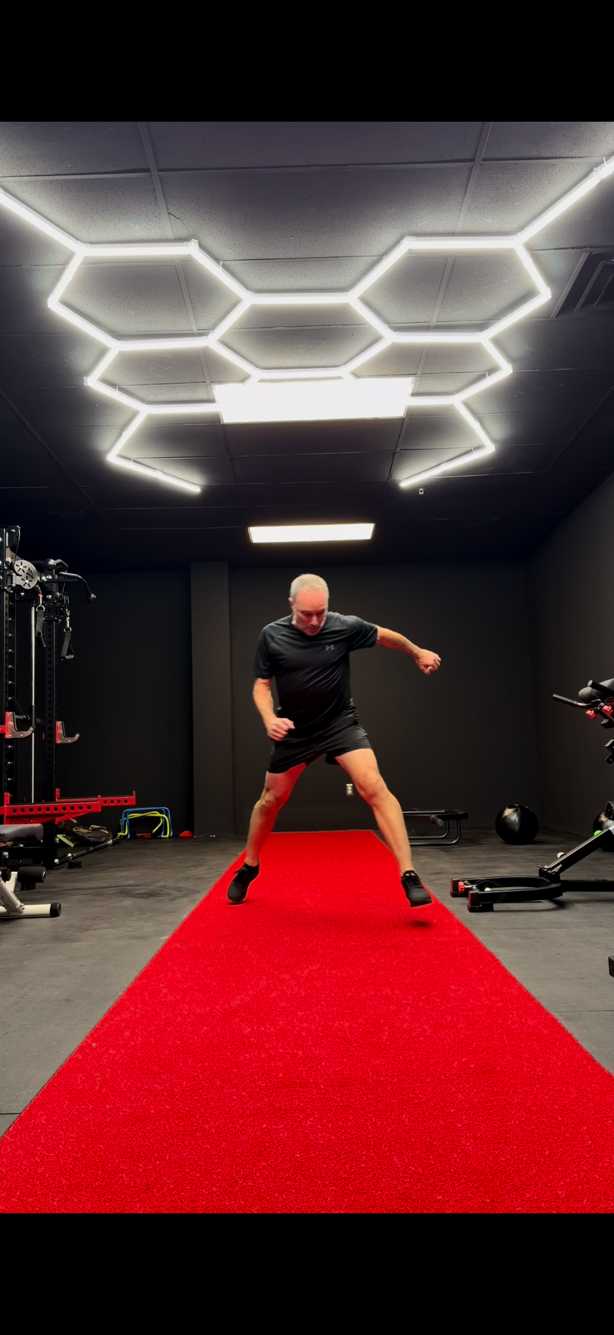 An older man in athletic clothing is jumping or landing on a red carpet in a gym, with workout equipment and black walls and ceiling with hexagonal light fixtures.