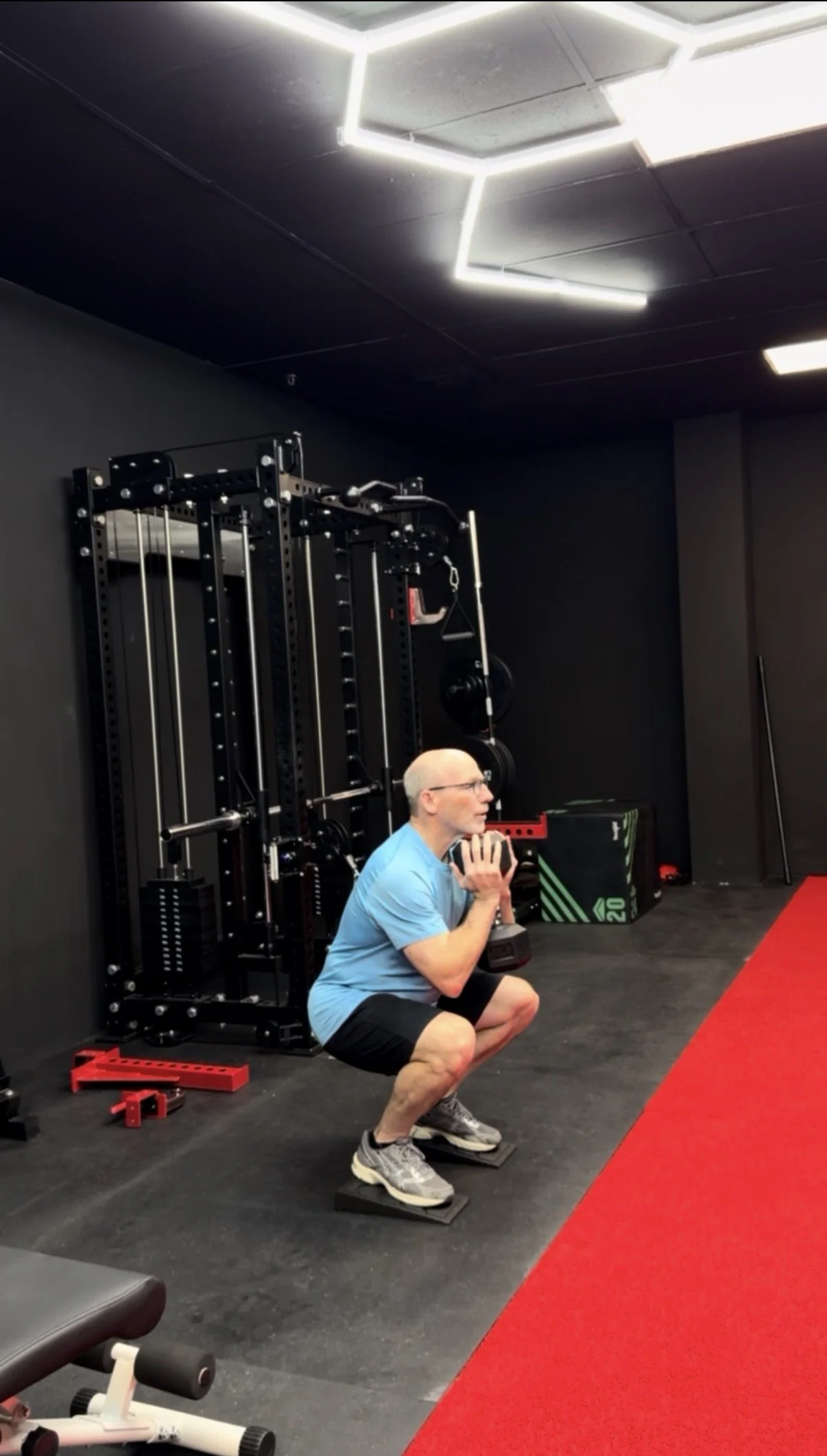 An older man doing a squat exercise while holding weights in a gym. He is positioned near black gym equipment and a red carpeted area.