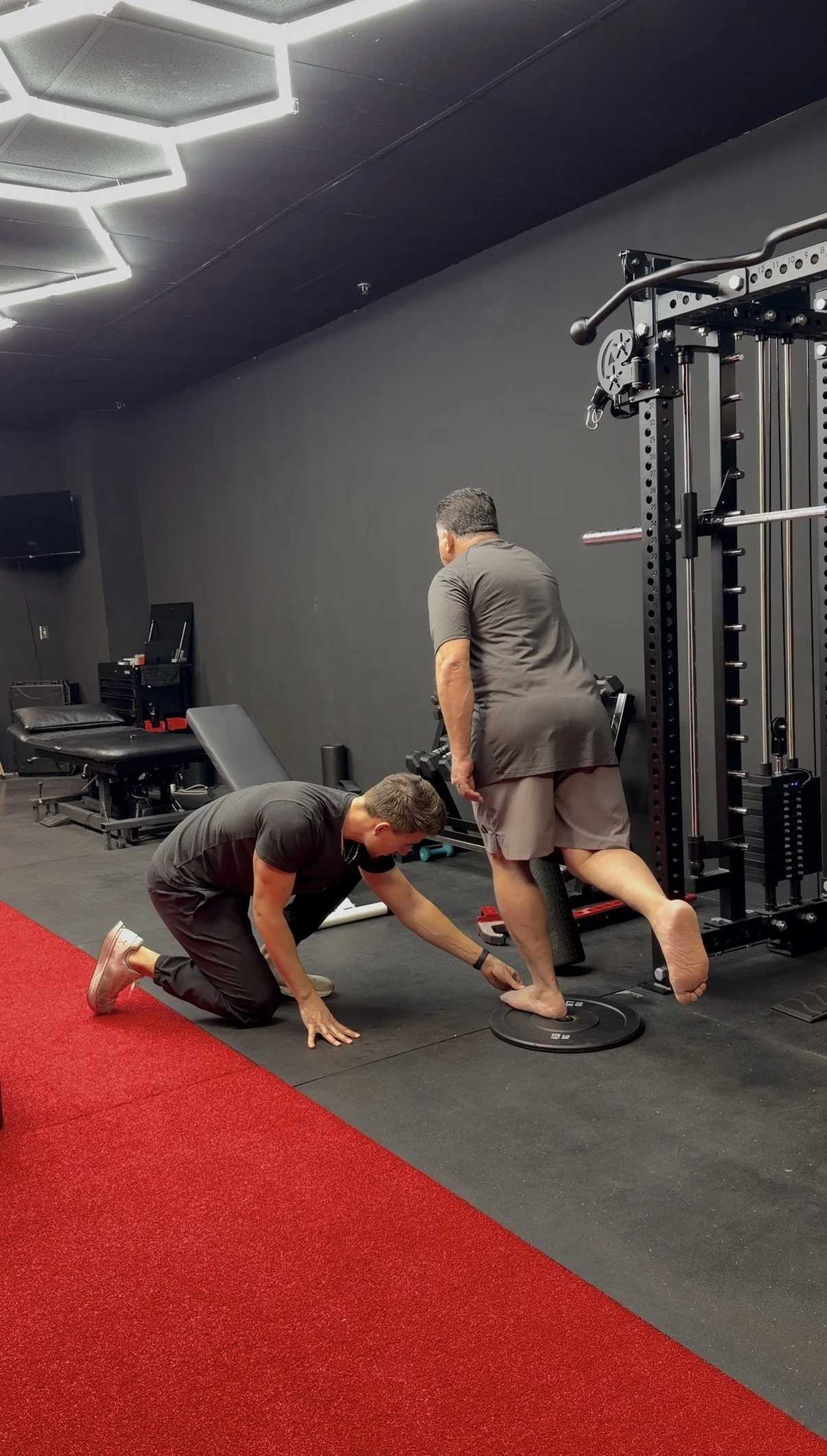 A trainer assisting a person with balance in a gym, with gym equipment and a red carpet in the background.