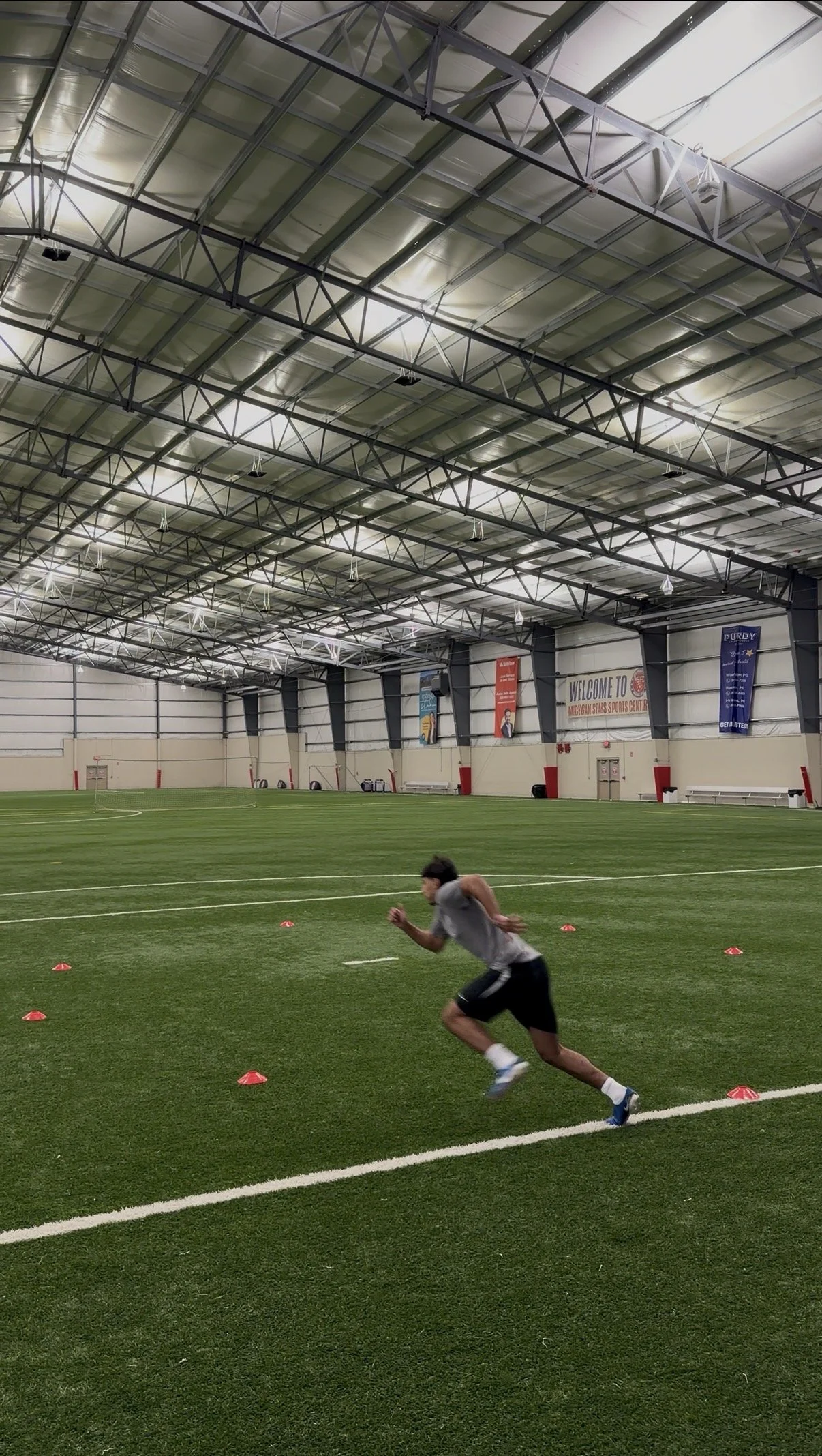 A person running in an indoor sports facility, with orange cones laid out in a pattern on the artificial turf field.