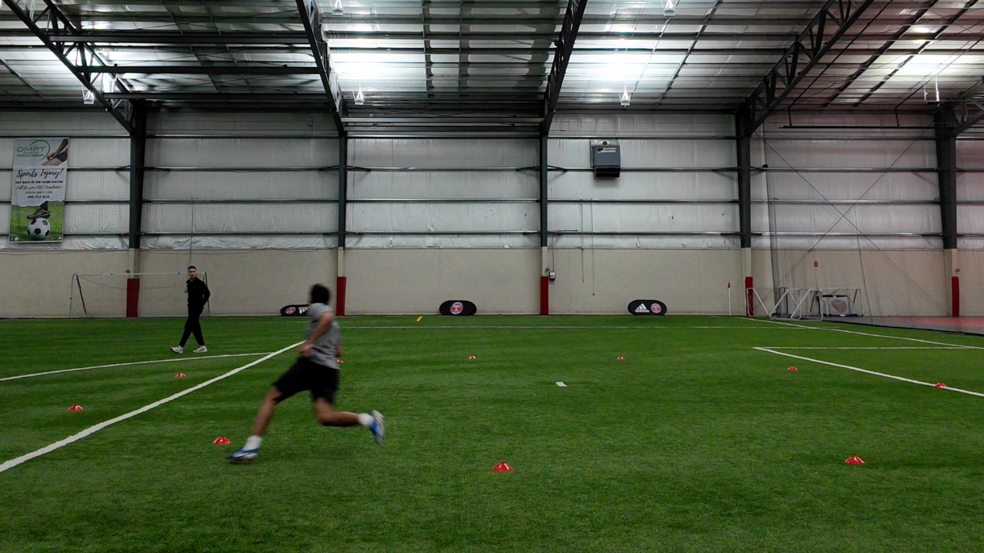 Indoor sports training facility with artificial turf, soccer goals, and agility cones set up for drills, with two individuals practicing.